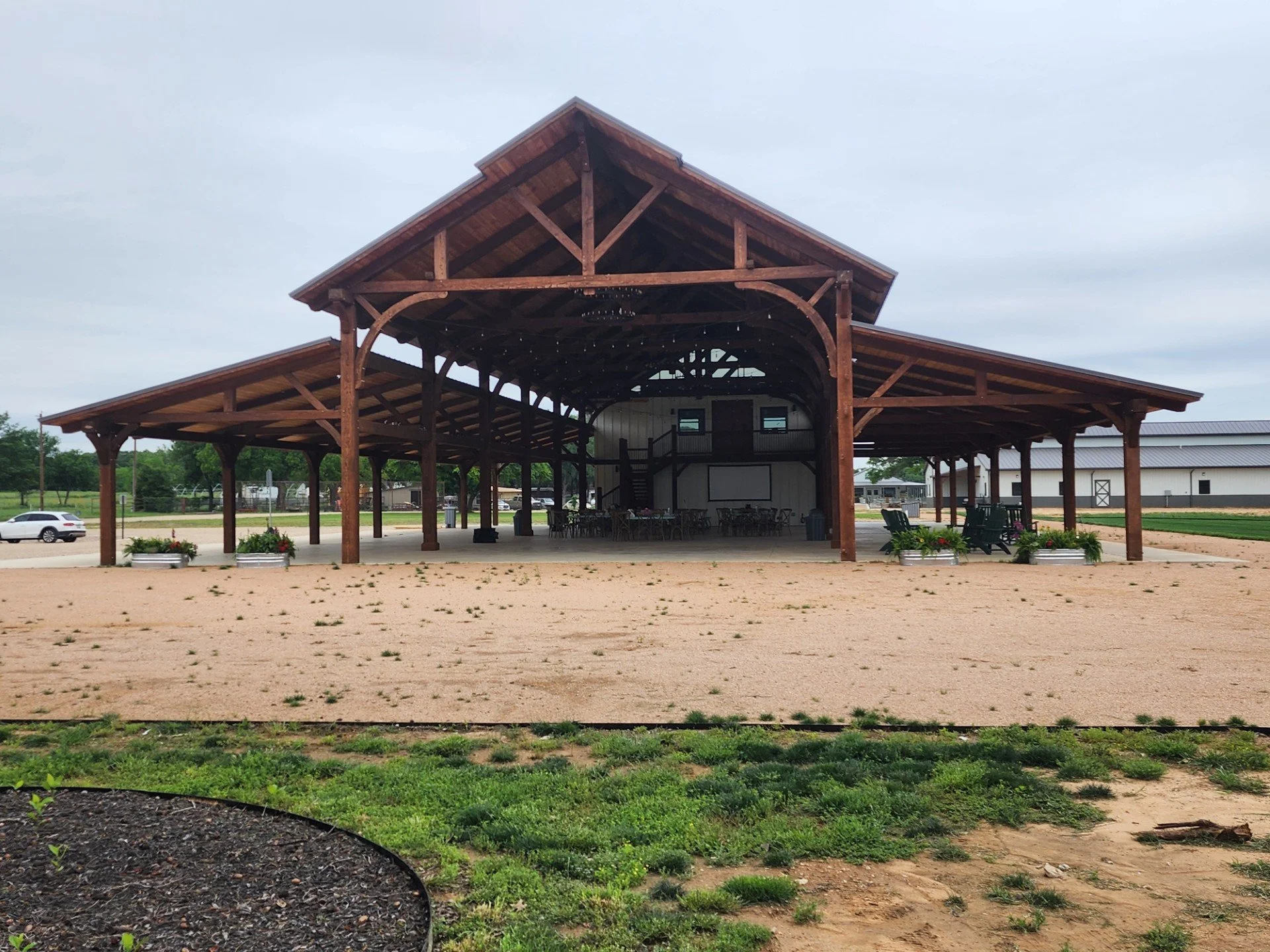 A wooden pavilion with a peaked roof and open sides in a rural area, with seating and a basketball hoop inside, surrounded by a landscape with grass, dirt, and parked cars.