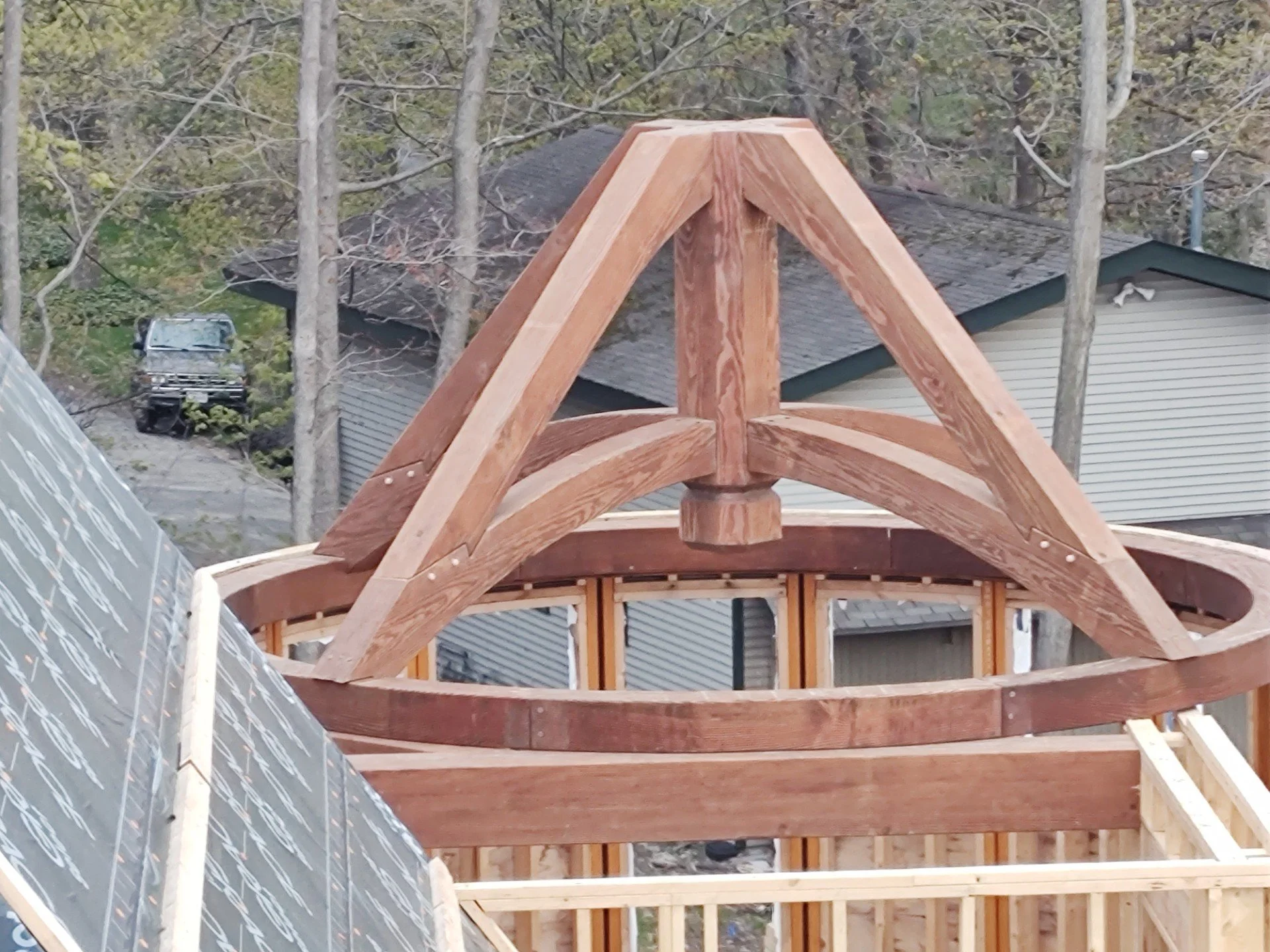 Under construction wooden roof frame on a building, with trees and a house in the background.