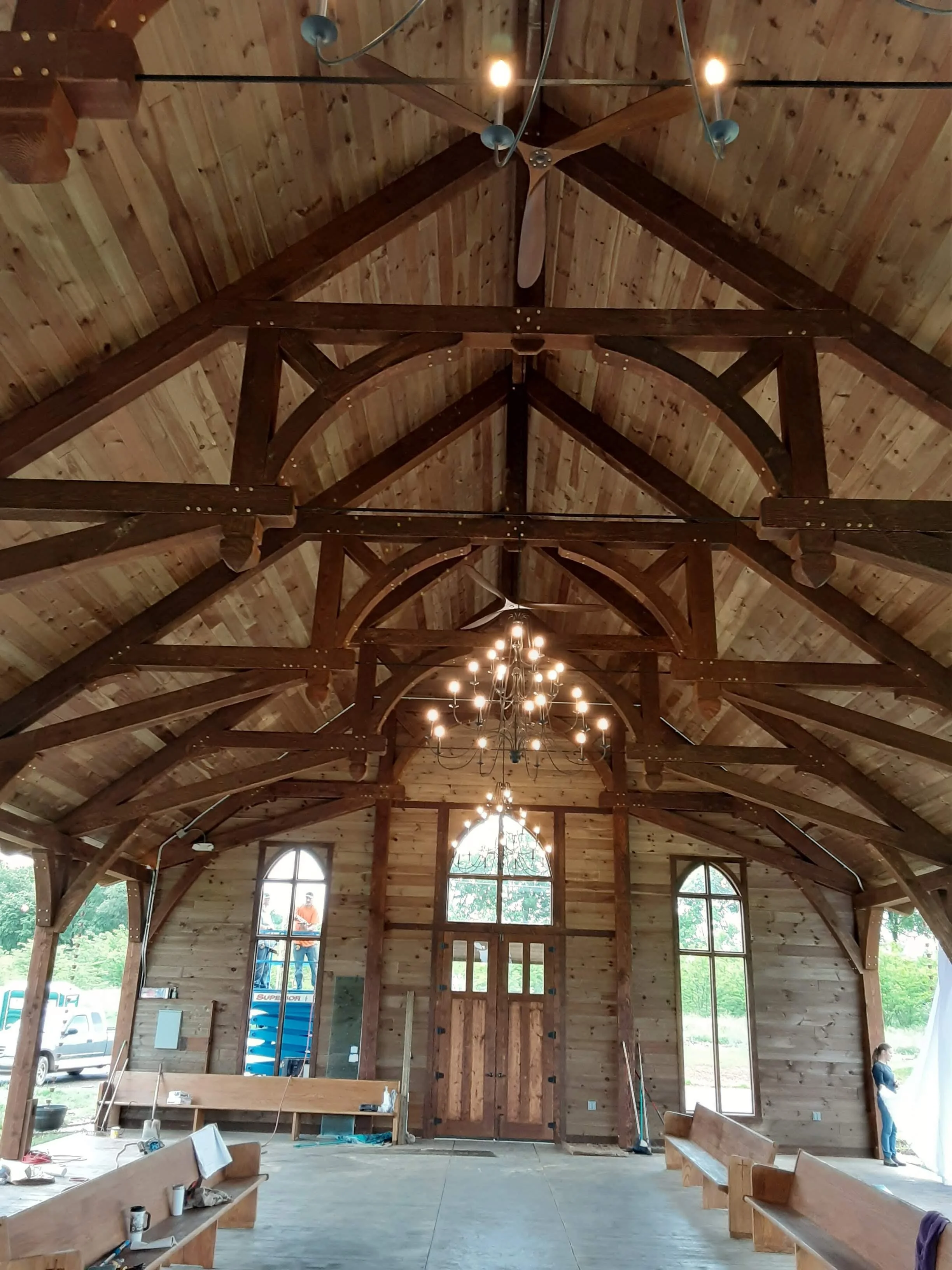 Interior of a wooden church under construction with exposed beams, chandeliers, and large windows, with a woman standing near the window and construction tools on the floor.