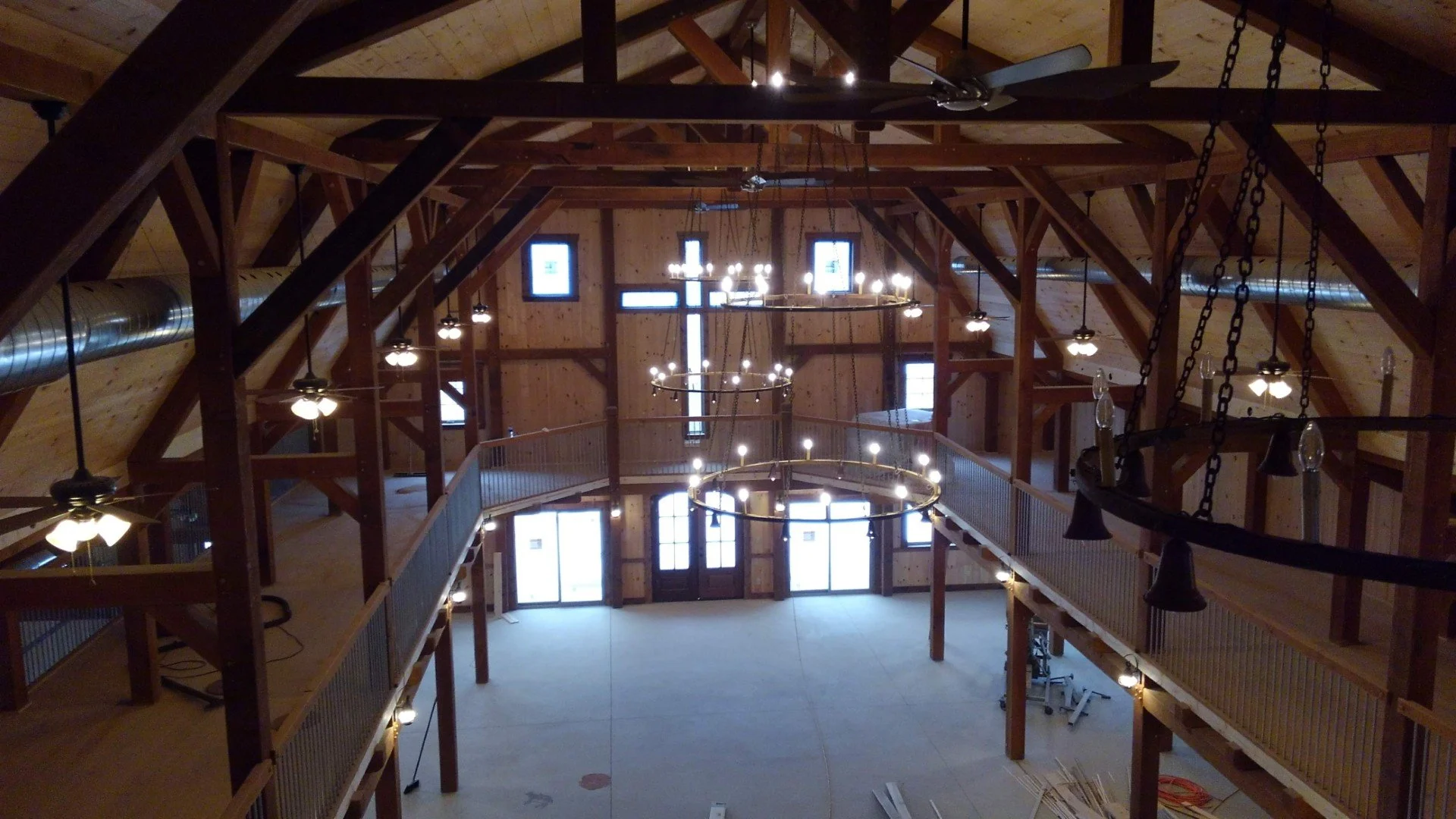Interior of a wooden church or hall with high ceiling, multiple chandeliers, fans, and large windows, viewed from an upper balcony.