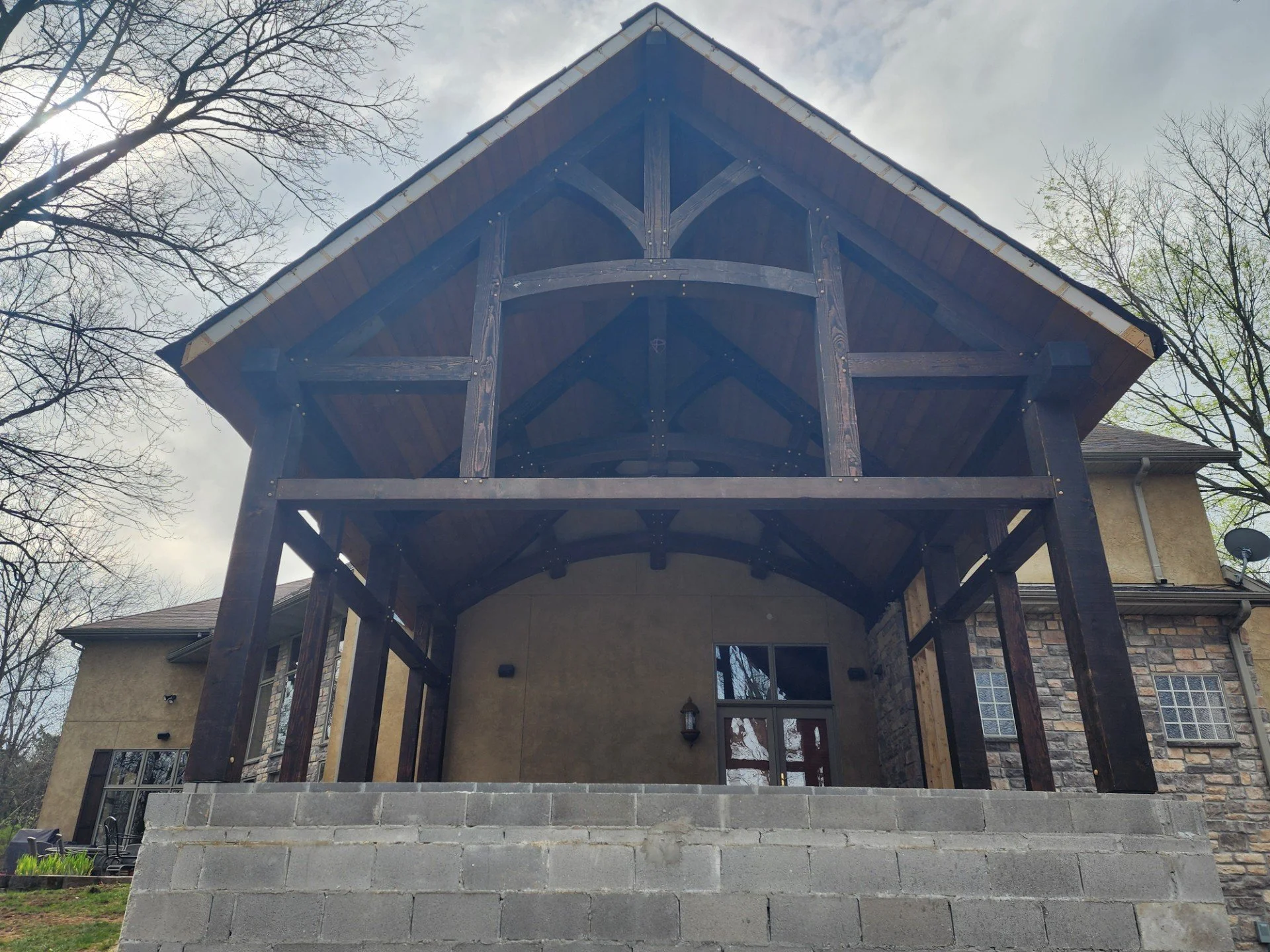 View from the ground showing a house with a large wooden balcony framework under construction, with brick and stone exterior walls, double-pane windows, and leafless trees in the background.