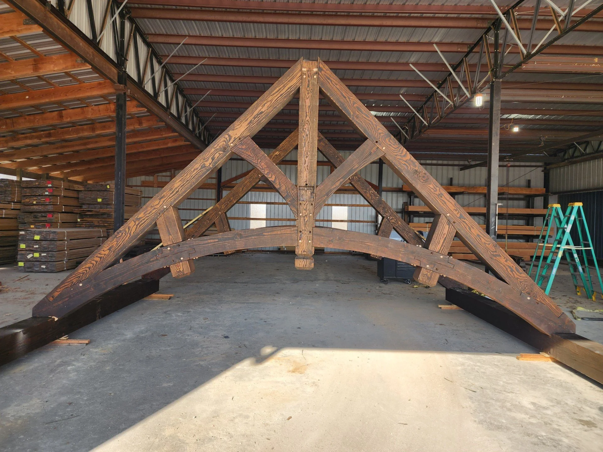 A wooden bridge under construction inside a large industrial warehouse, with stacks of wood on the side and a green ladder on the right, and metal roofing overhead.
