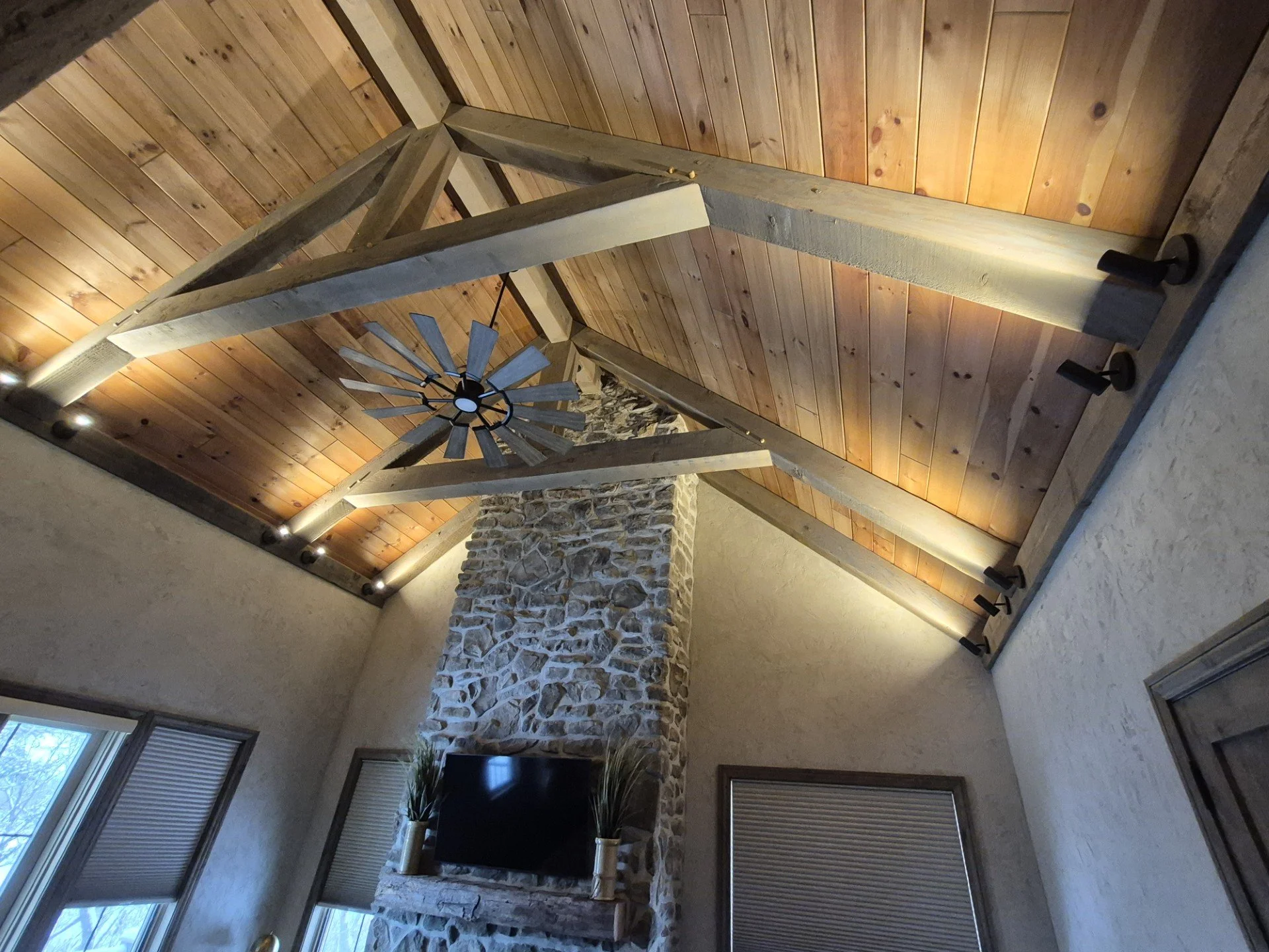 Interior of a room with a wooden, high, vaulted ceiling, a stone fireplace, windows with blinds, and a chandelier with wooden blades.