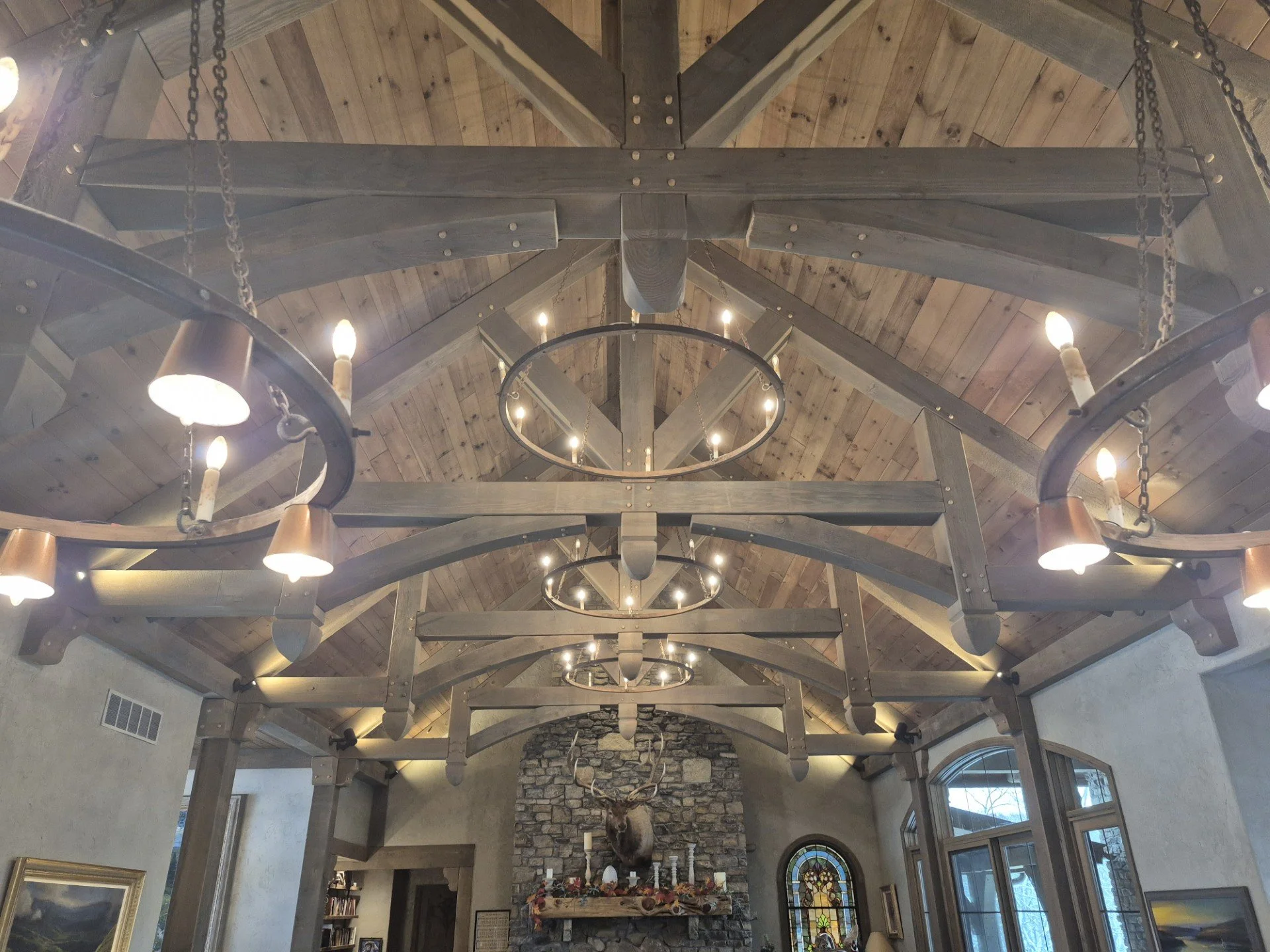 Interior of a rustic room with a high, wooden vaulted ceiling, chandeliers with candle-shaped lights, and a stone fireplace with a mounted deer head above it.