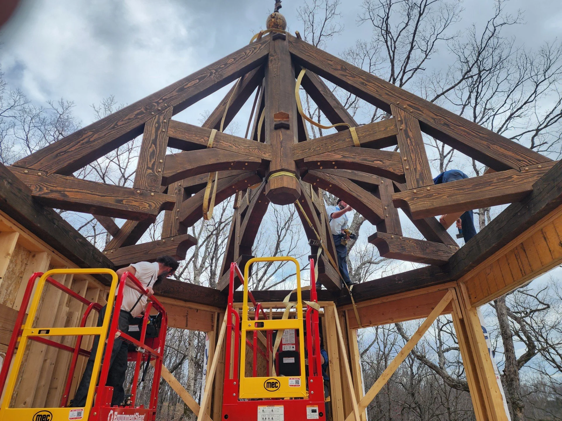 Workers constructing a wooden structure, possibly a tower or a treehouse, using a red and yellow lift with MEC branding. The structure has a large wooden framework with arched beams and is set outdoors among leafless trees on a cloudy day.