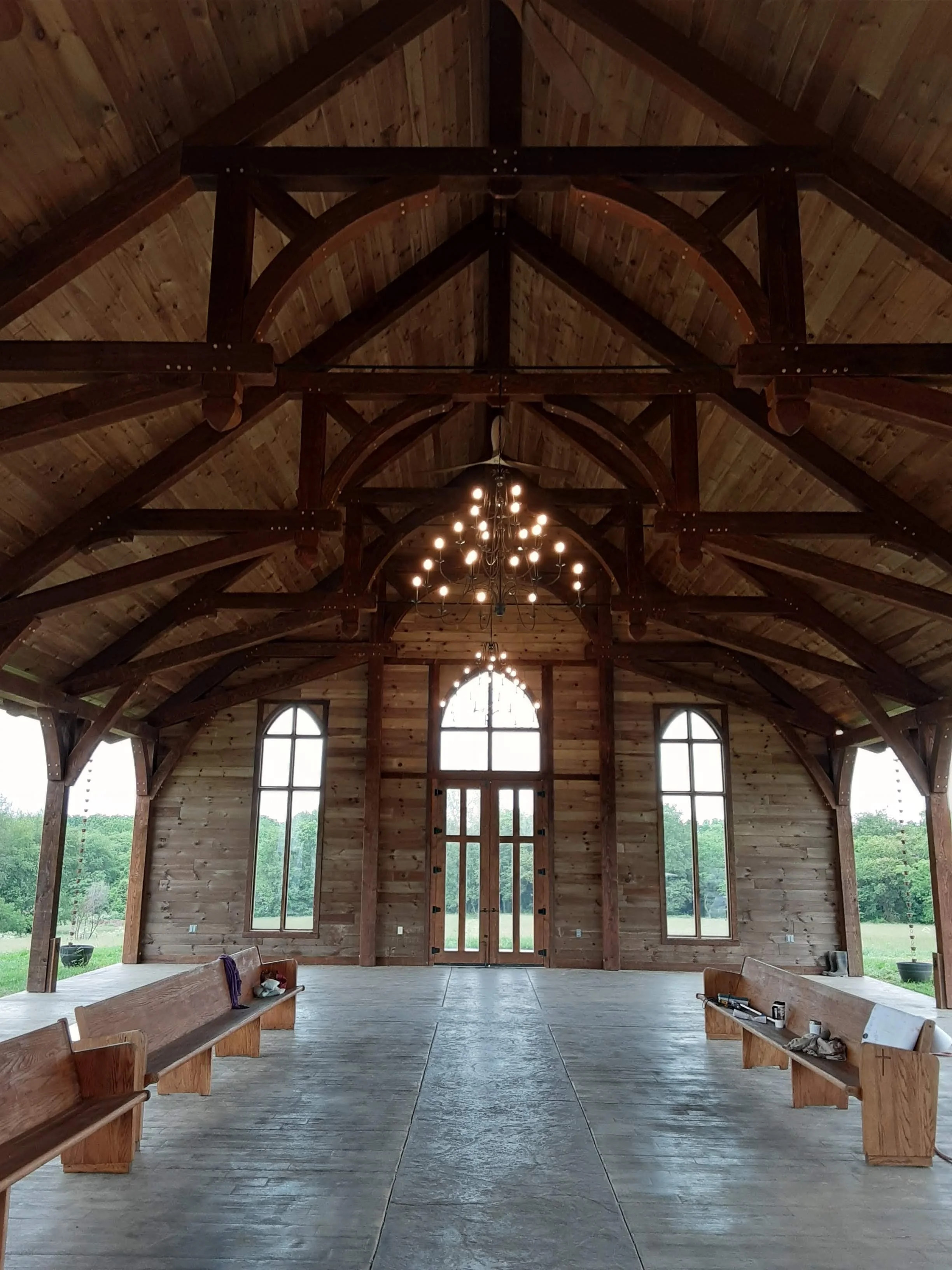 The interior of a wooden church or chapel with high vaulted wooden ceiling, hanging chandelier, large arched windows, wooden pews on either side, and open double doors at the back with scenic view outside.
