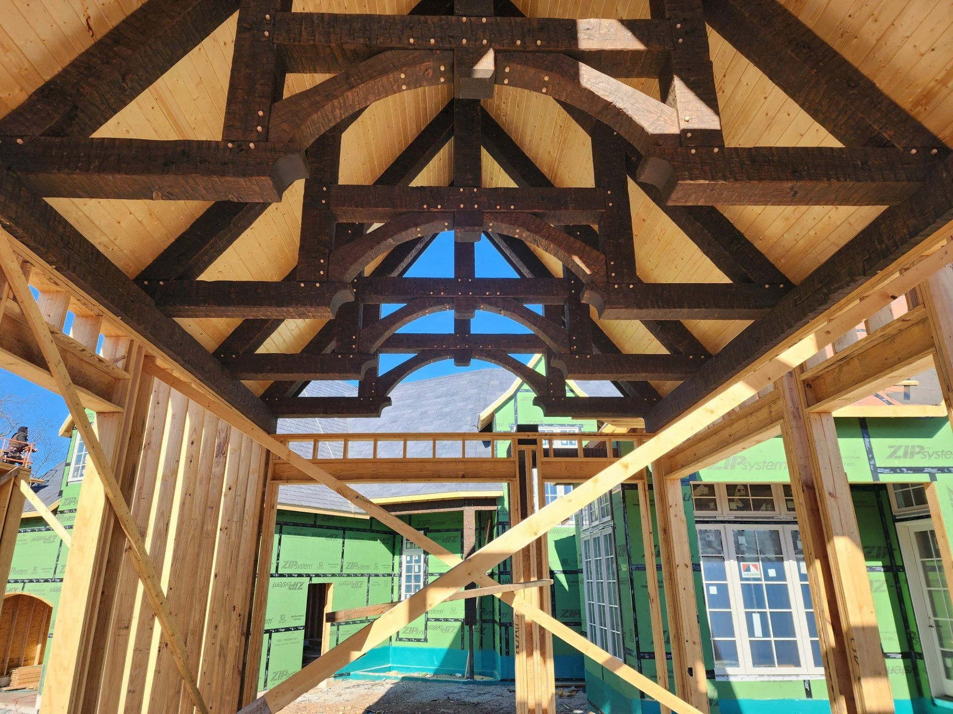 Wooden framing and beams of a house under construction, with green sheathing and a window visible in the background, and blue sky overhead.