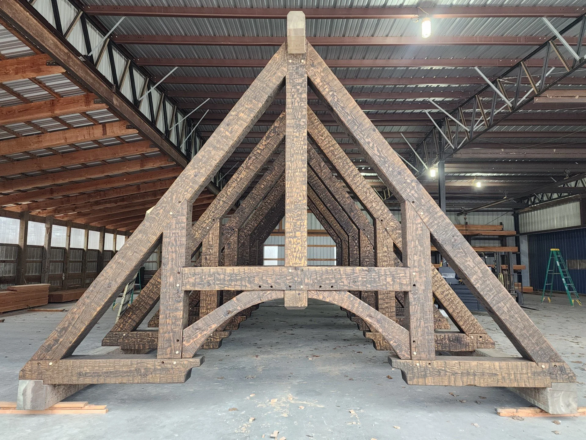 A wooden A-frame structure being built inside a warehouse with metal walls and ceiling, scaffolding, and construction materials nearby.
