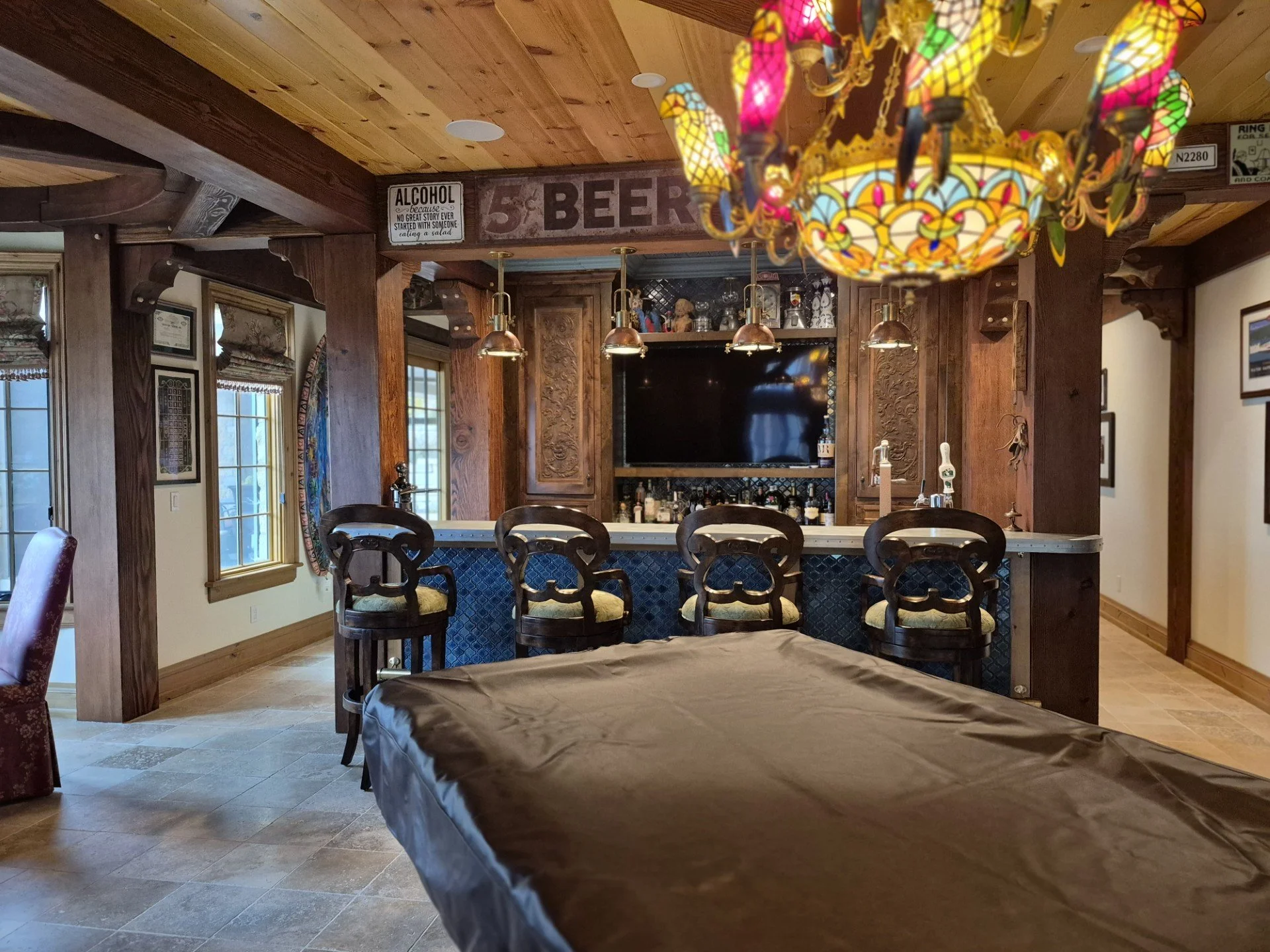 A cozy bar area with a wooden counter, surrounded by four wooden chairs with cushioned seats. The bar is decorated with carved wood panels, and various bottles and glassware are displayed behind it. Above the bar, there are three copper pendant light