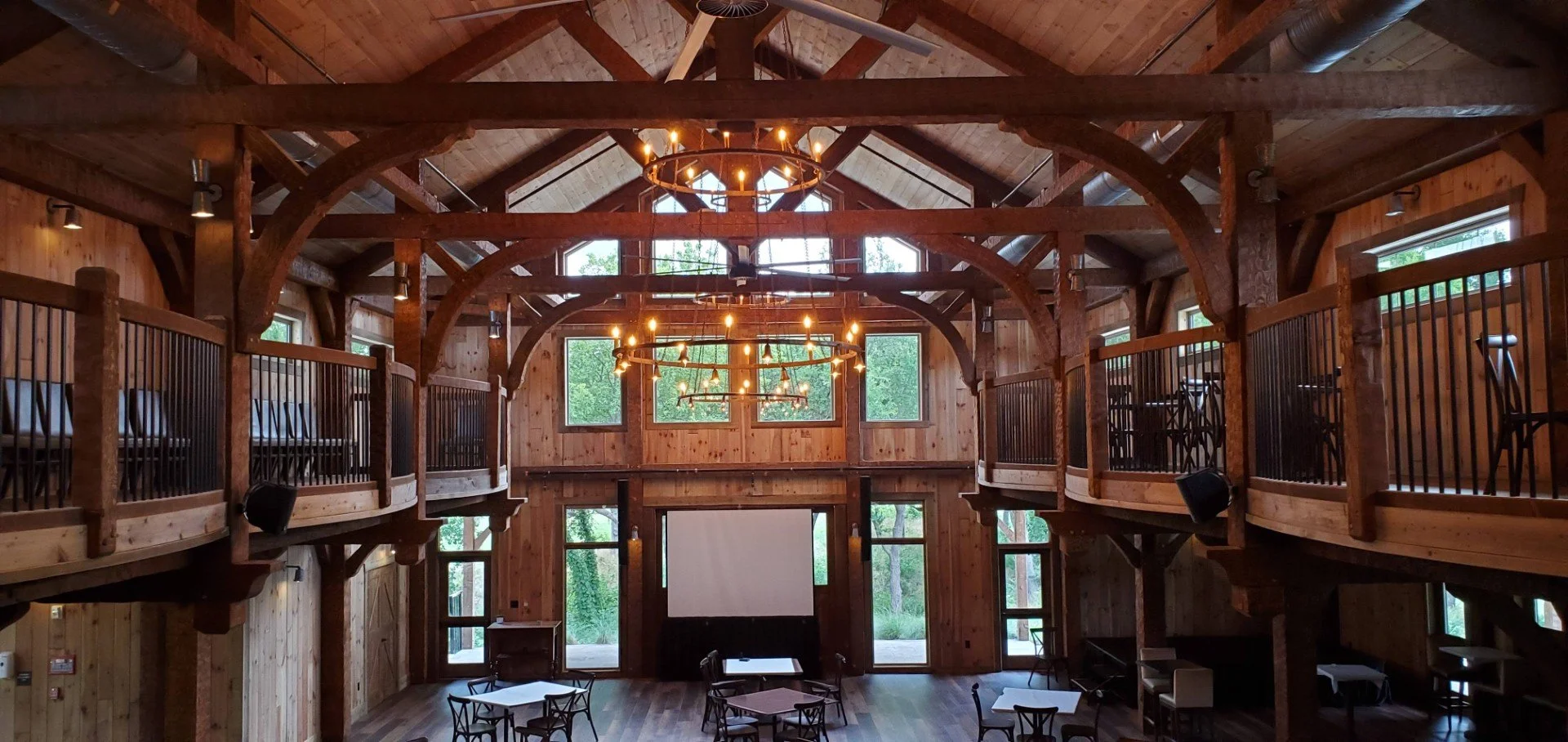 Interior of a rustic wooden lodge with high vaulted ceilings, multiple chandeliers, and an open second-floor balcony with seating.