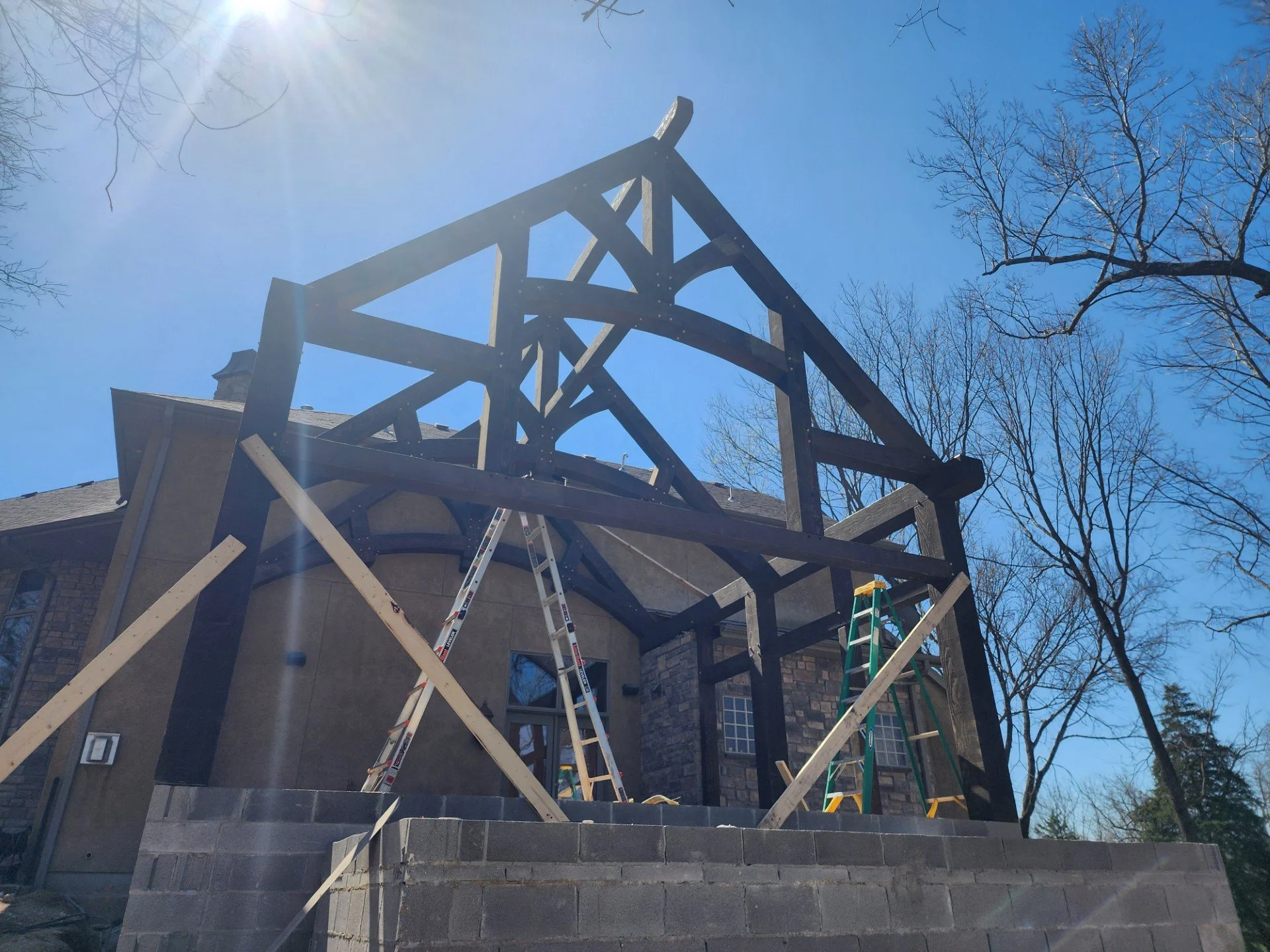 Construction site with a house frame being built, ladders, and scaffolding under a clear blue sky with sunlight and leafless trees.