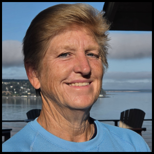 A woman with short blonde hair smiling outdoors near water with boats and mountains in the background.