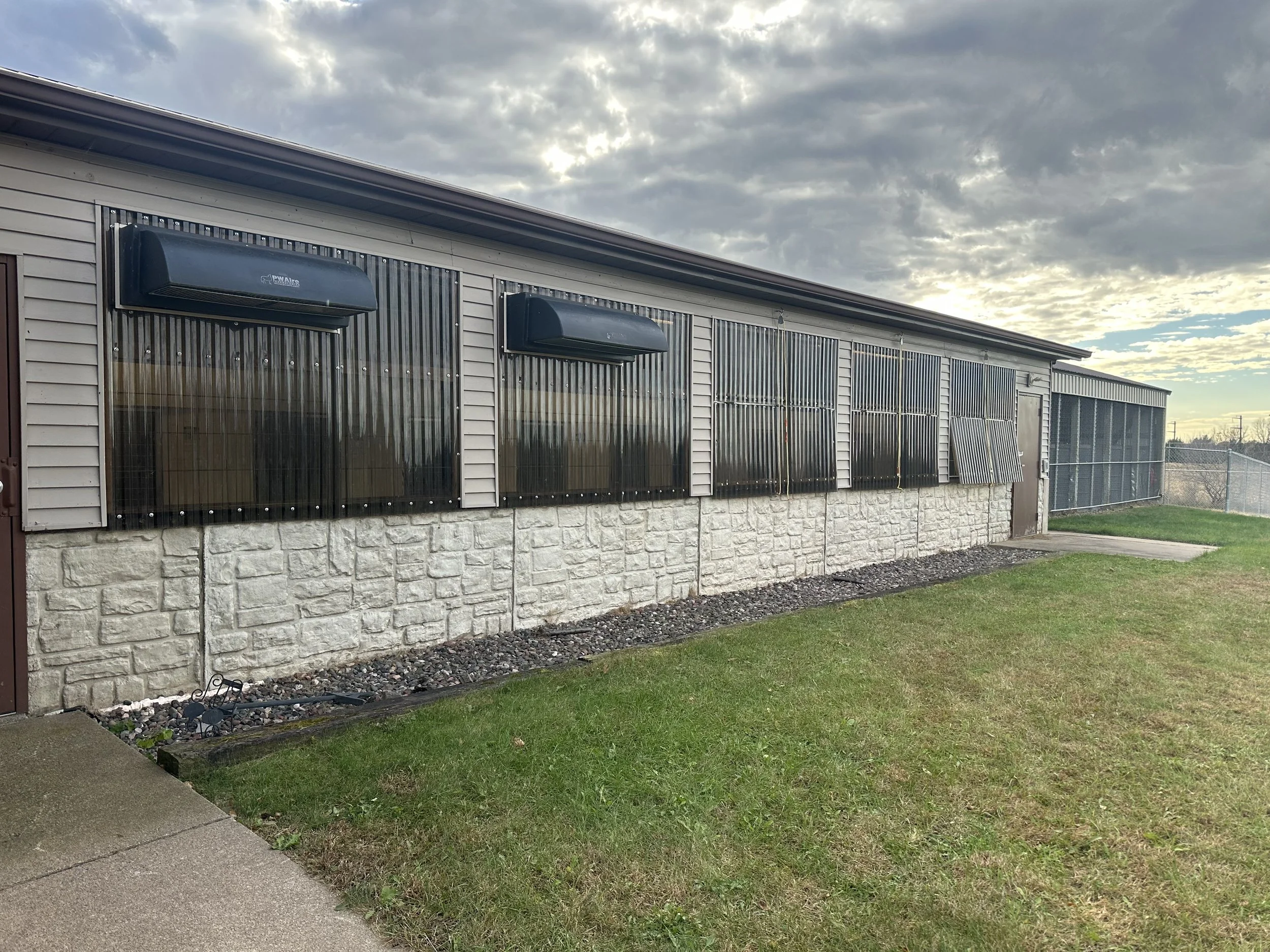 Exterior view of a building with vertically aligned solar window covers, a stone foundation, and a grassy lawn, under a clouded sky.
