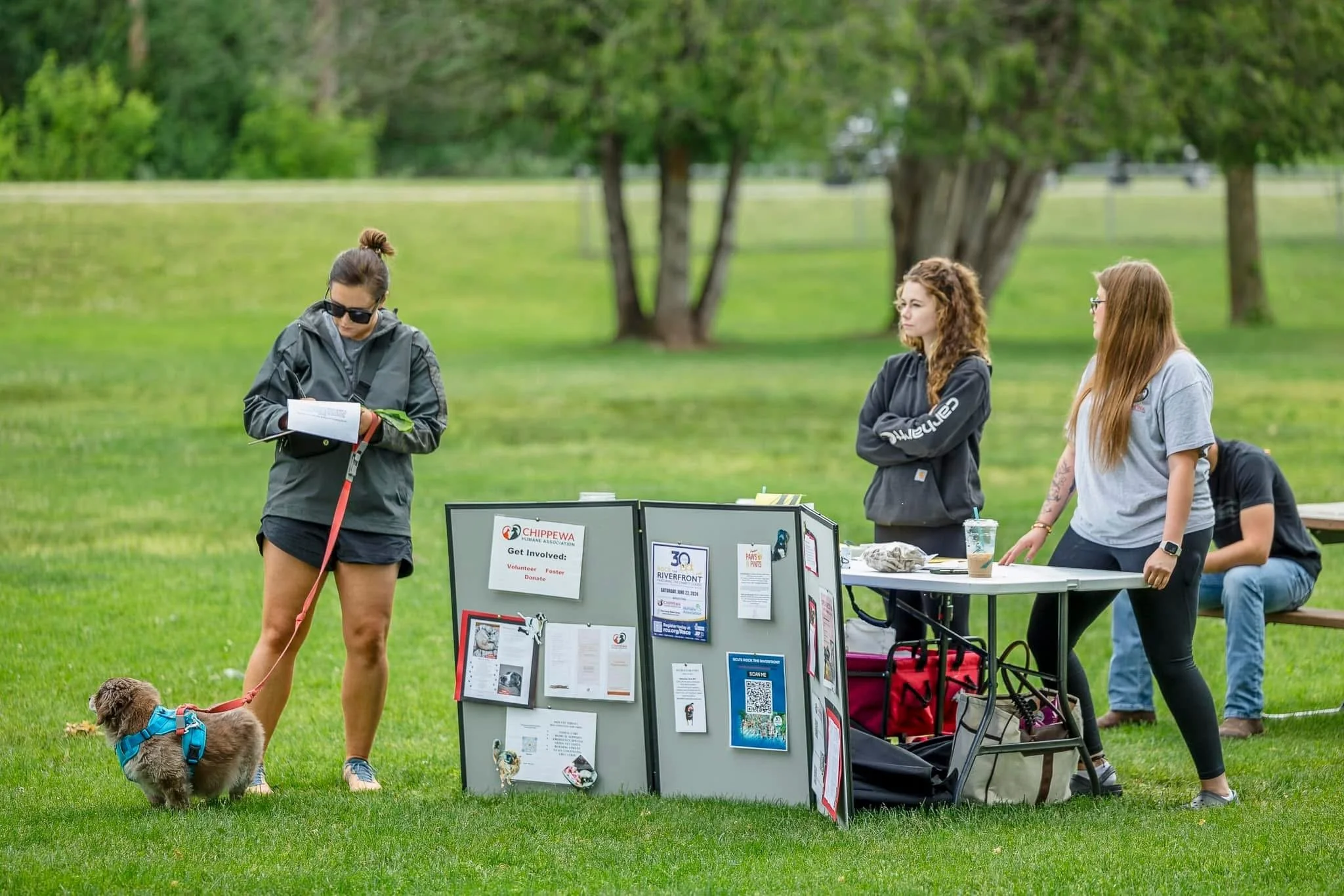 A woman in black shorts and sunglasses stands with a small dog on a leash at an outdoor event table. Two women with long hair, wearing casual shirts, stand nearby at the event table, which has various flyers and signs. A man is seated at the table in the background, behind a bench, surrounded by green grass and trees.