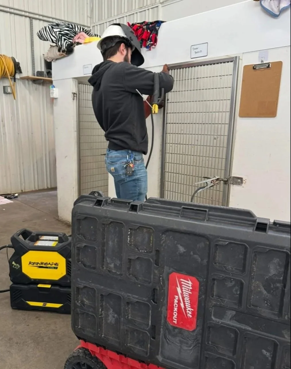A man wearing a helmet is working with a power drill inside a fenced enclosure marked 'Dog Yard.' There is a toolkit labeled 'Milwaukee PACKOUT' on the floor and some clothing or fabric items on a shelf overhead.