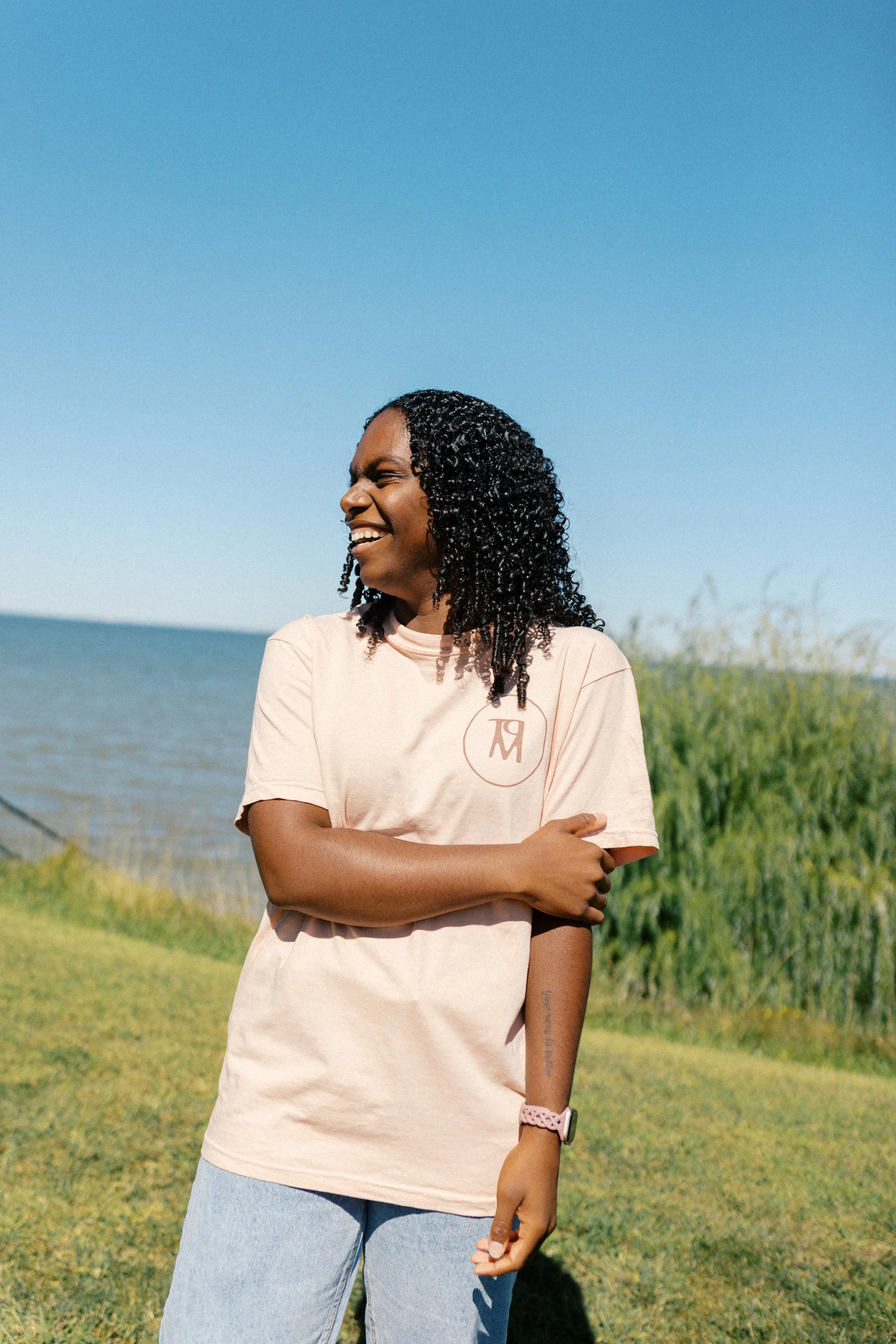 A woman with curly black hair smiling and looking away outdoors near a body of water, wearing a pastel pink t-shirt and light blue jeans.