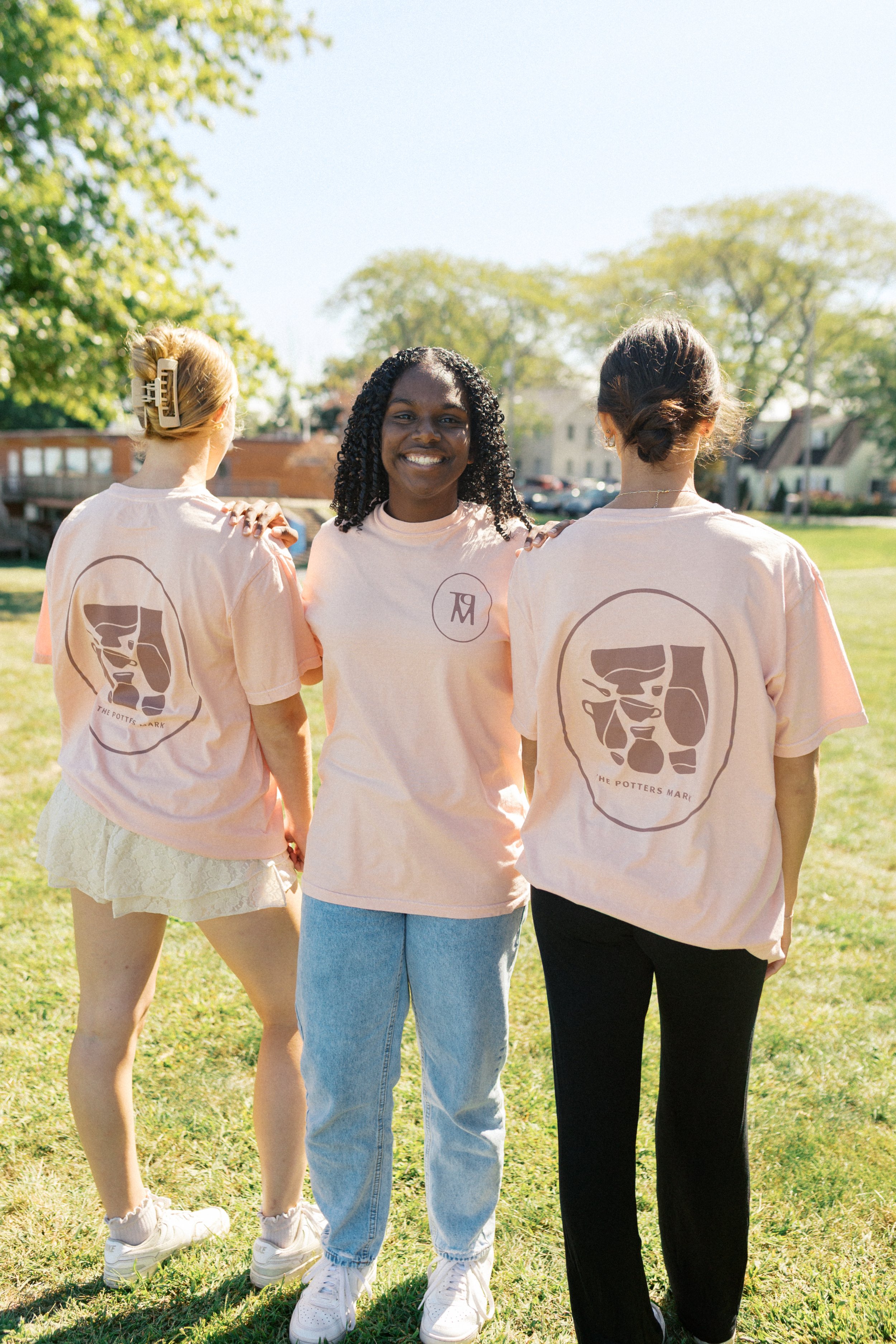 Three women standing outdoors on a sunny day, wearing matching light pink T-shirts with a pottery design and the text 'The Potters Mark.'