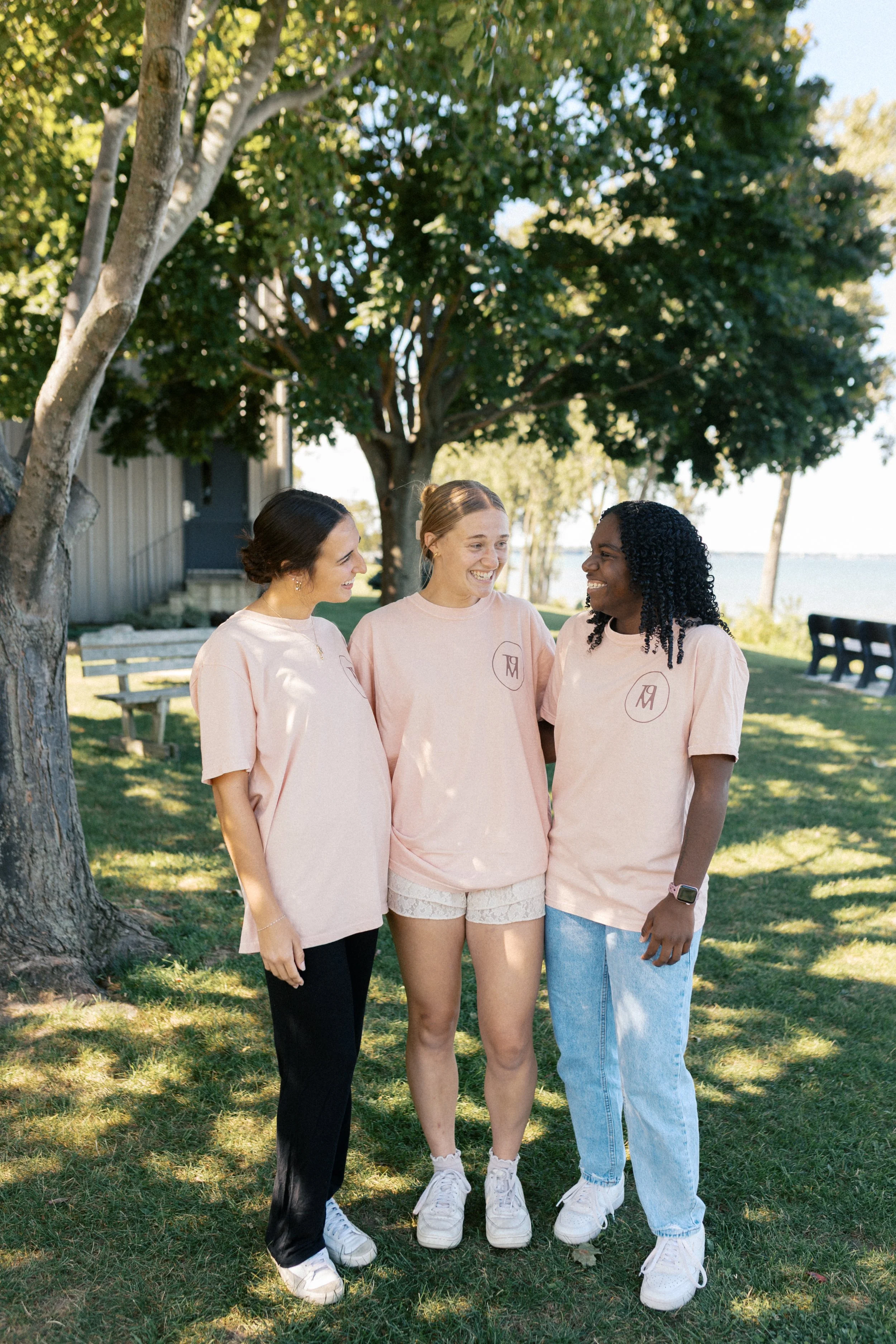 Three women standing close together outdoors under a tree, smiling and laughing, wearing matching pink T-shirts with a monogram logo.