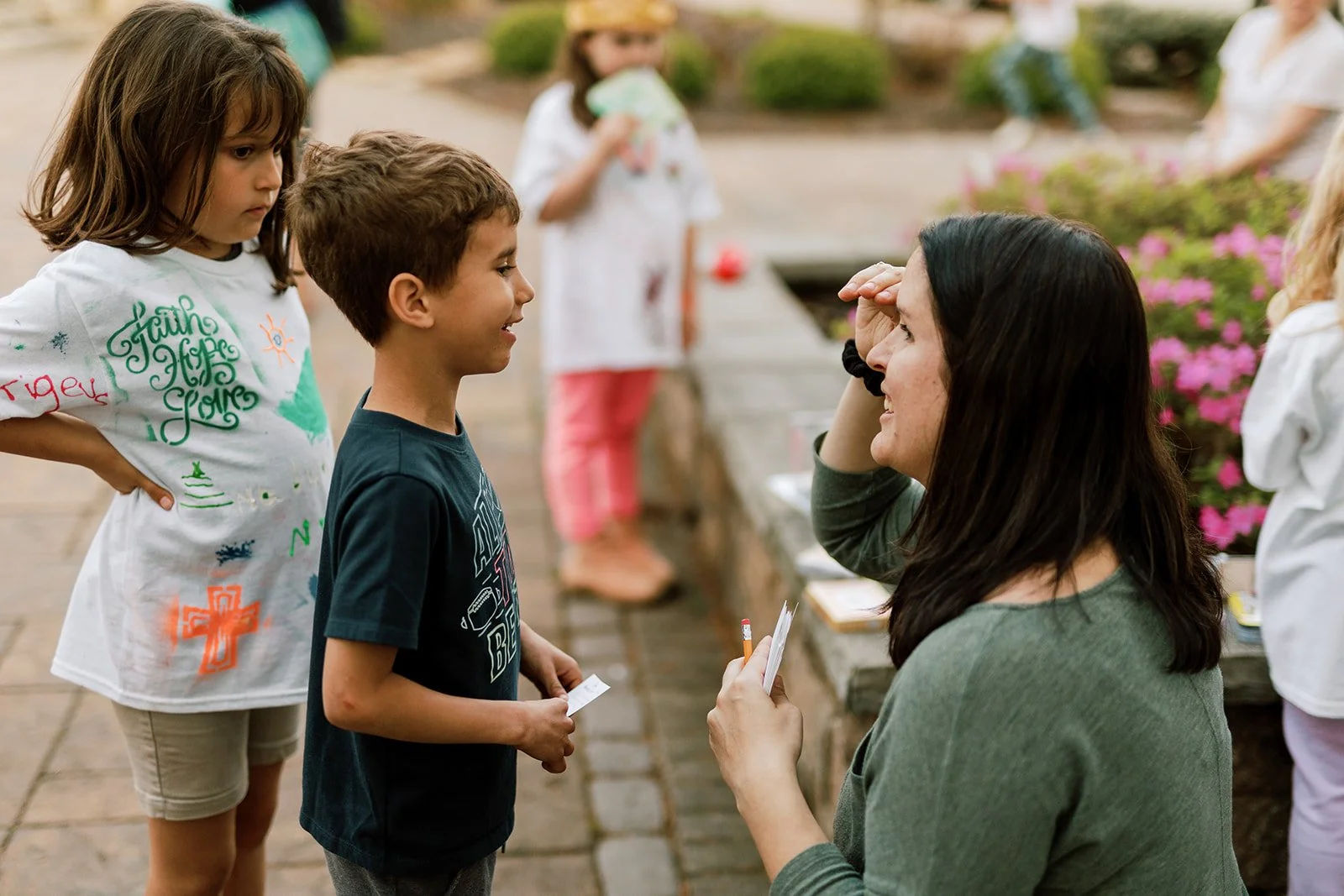 A woman smiling and talking to a boy outdoors, with children in the background, some holding papers or balloons, near pink flowers.