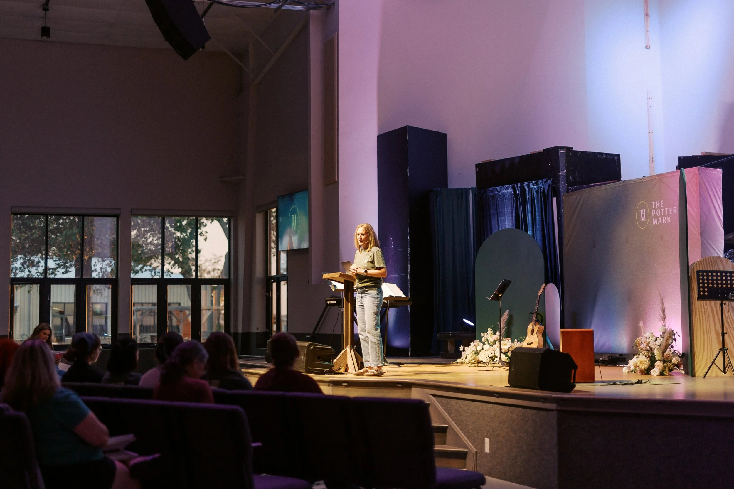 A woman speaking at a podium on stage with musical instruments and flowers, audience in seats, large windows showing trees outside.