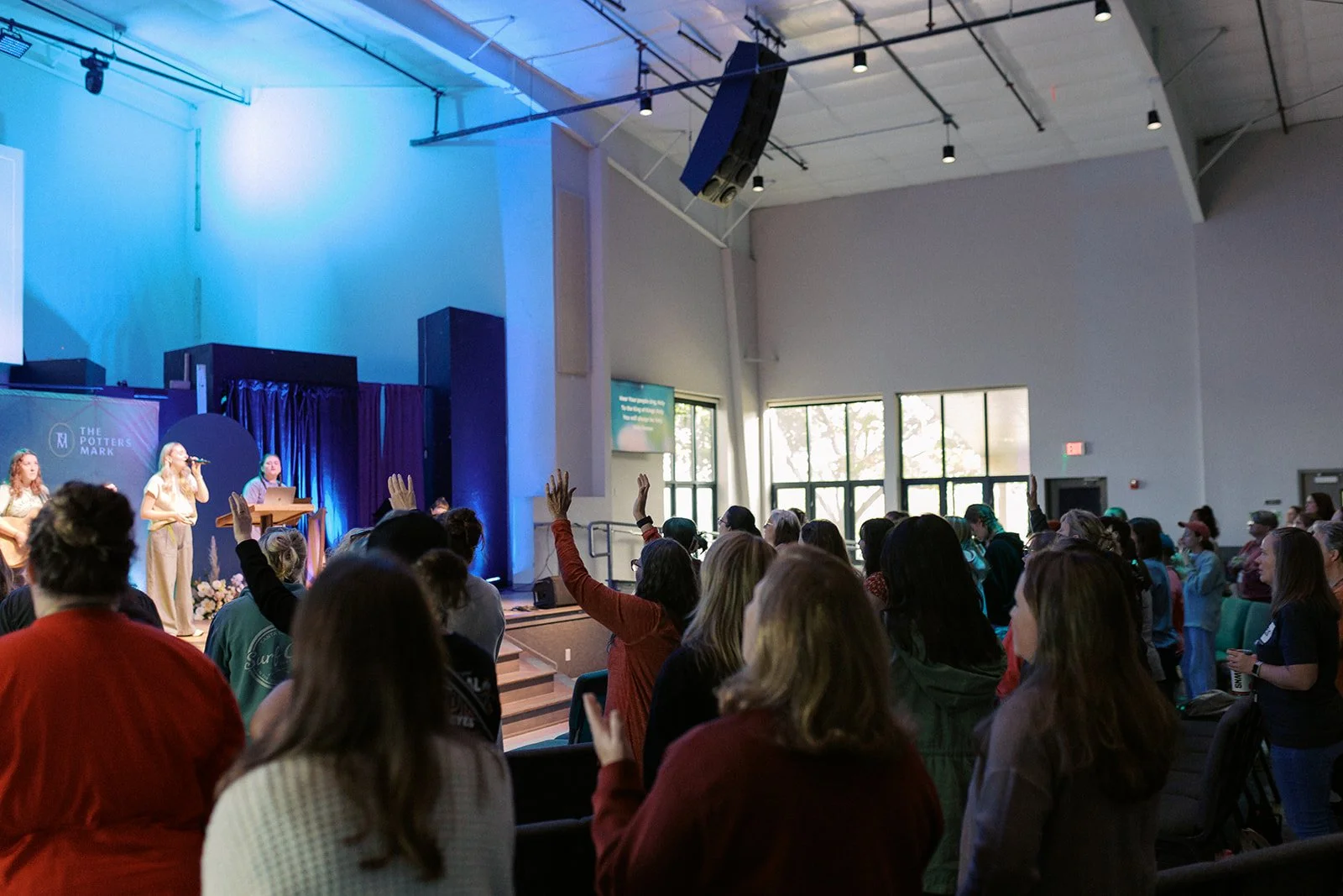 People gathered in a church or conference hall, participating in a group activity or worship, with some raising their hands, and a stage in the front where musicians and speakers perform.