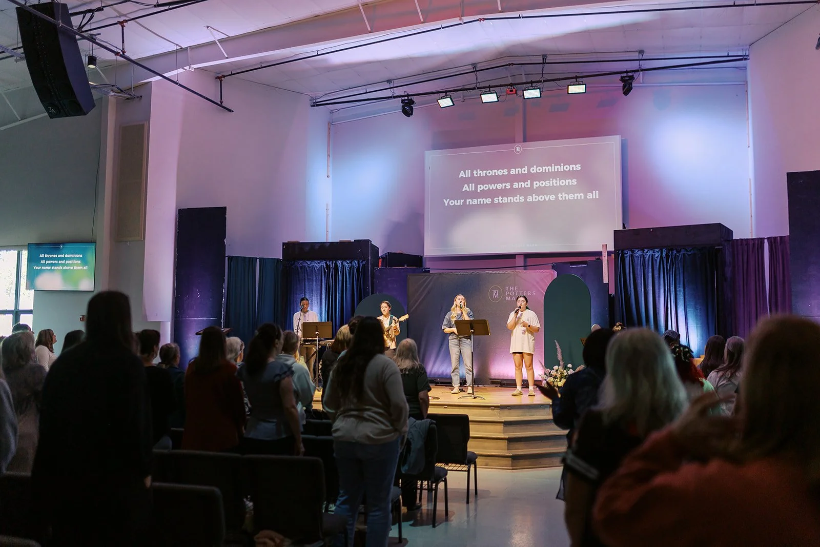 A church congregation gathered in a sanctuary with a stage where a band is performing. The band members are singing and playing instruments, and a song with lyrics is projected on a screen behind them. The audience is standing, some are holding flowe