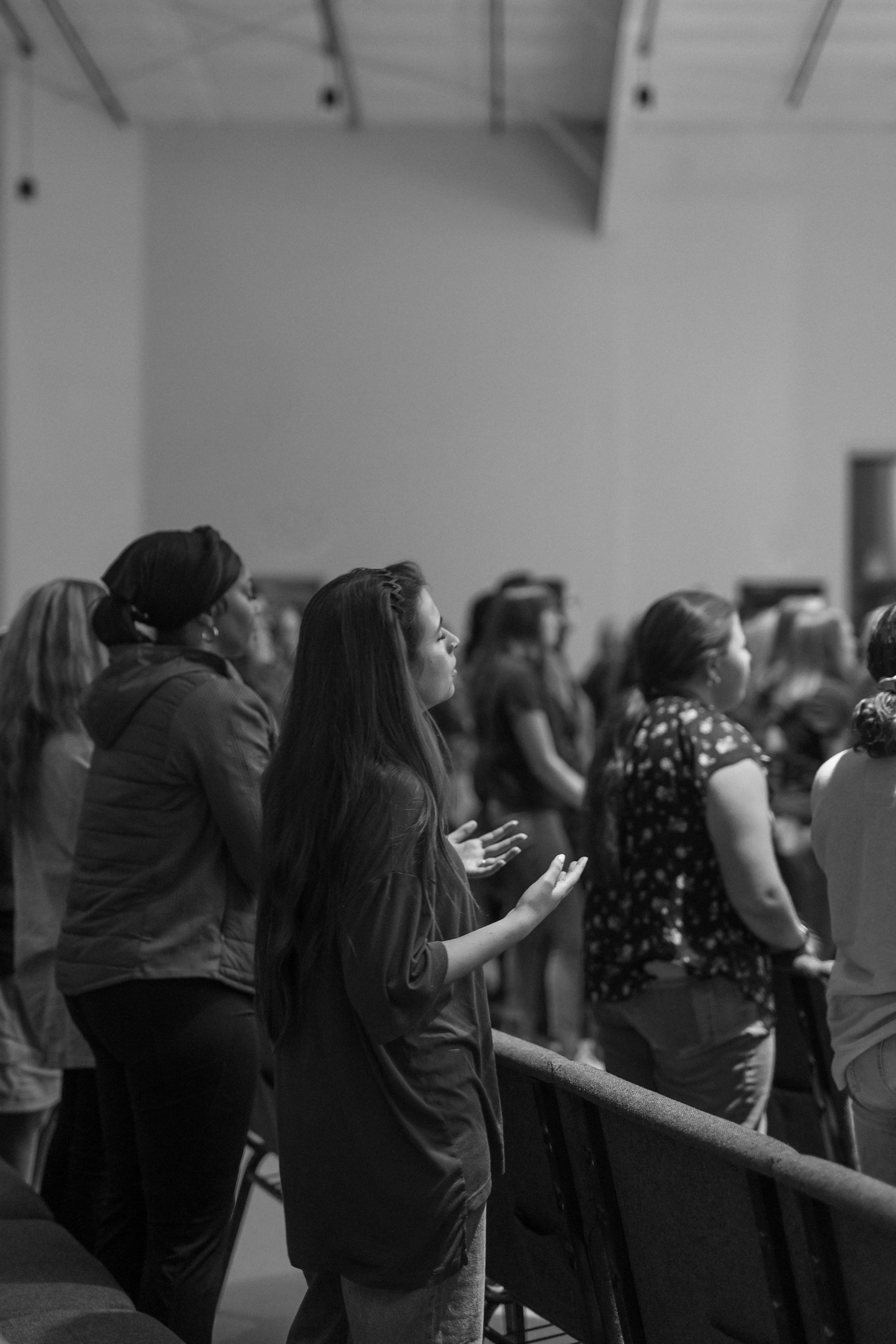 Black and white photo of women standing and praying in a church or auditorium. They have their eyes closed or looking upward with hands raised or clasped, indicating worship or prayer.
