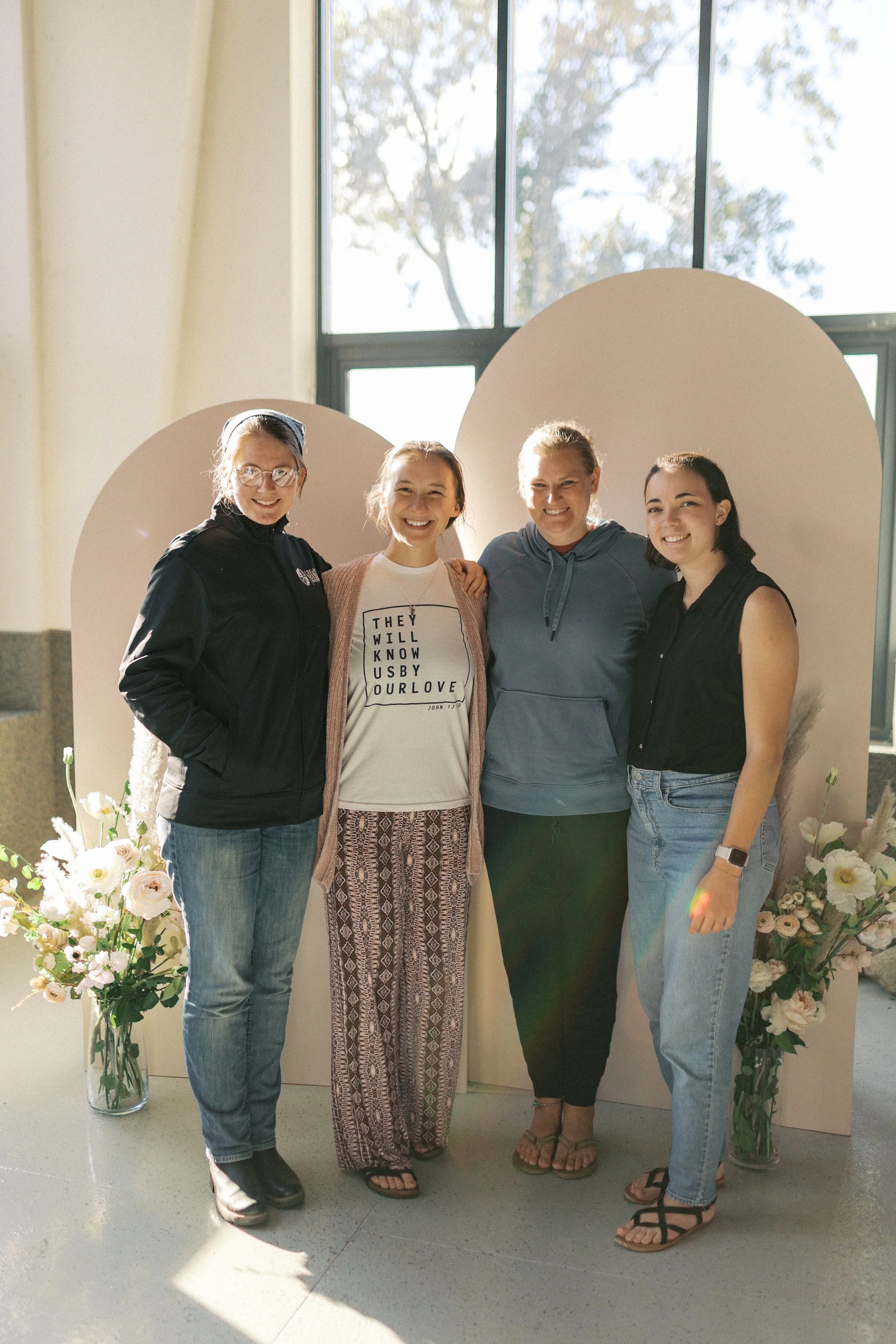 Four women standing together indoors, smiling for the camera, with large flower arrangements on either side, and a window behind them letting in natural light.