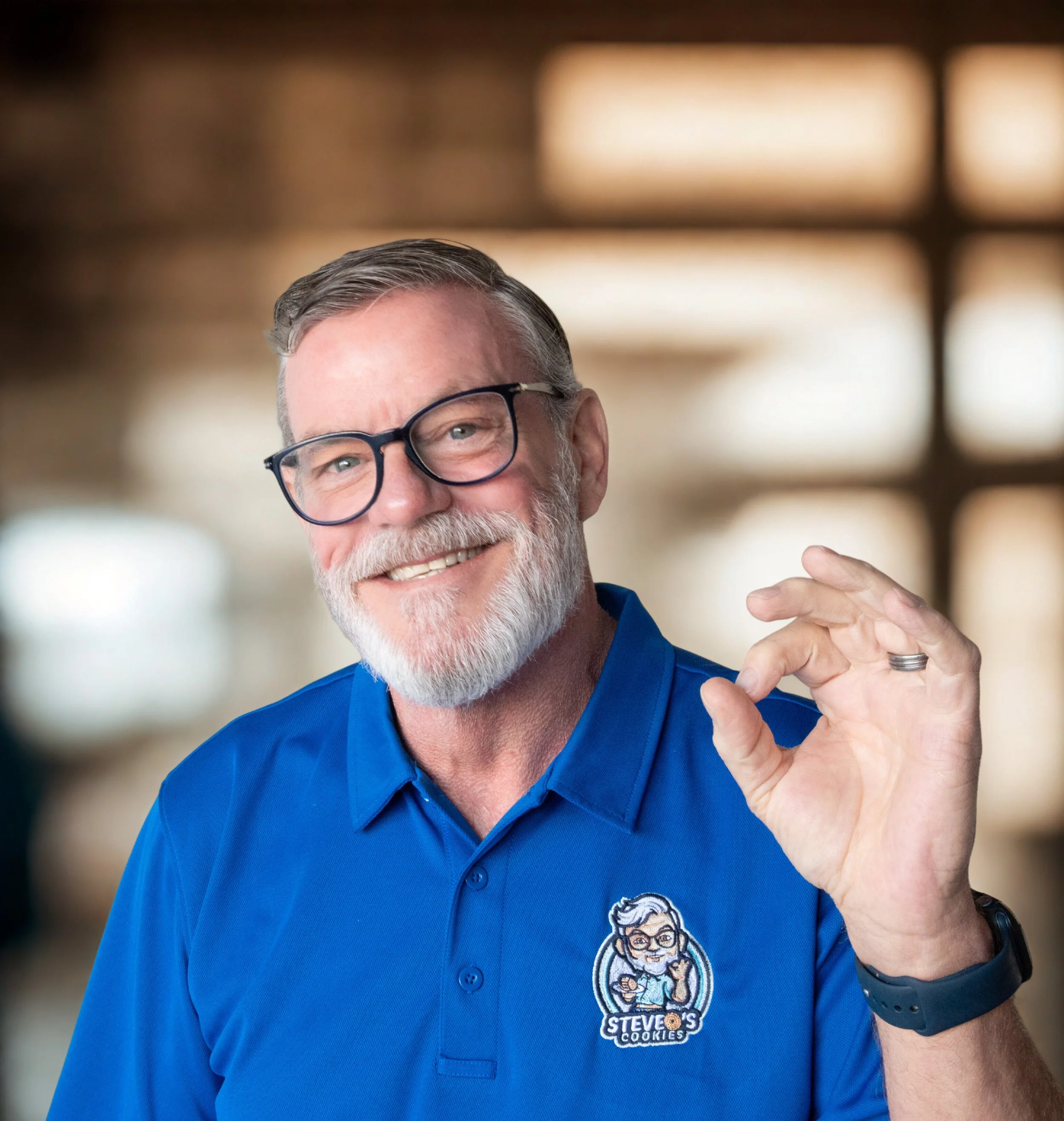 A smiling older man with gray hair, a beard, and glasses, wearing a blue polo shirt with a logo that says "Steve's Cookies" and a cartoon image of a man with glasses. He is making an OK gesture with his right hand.