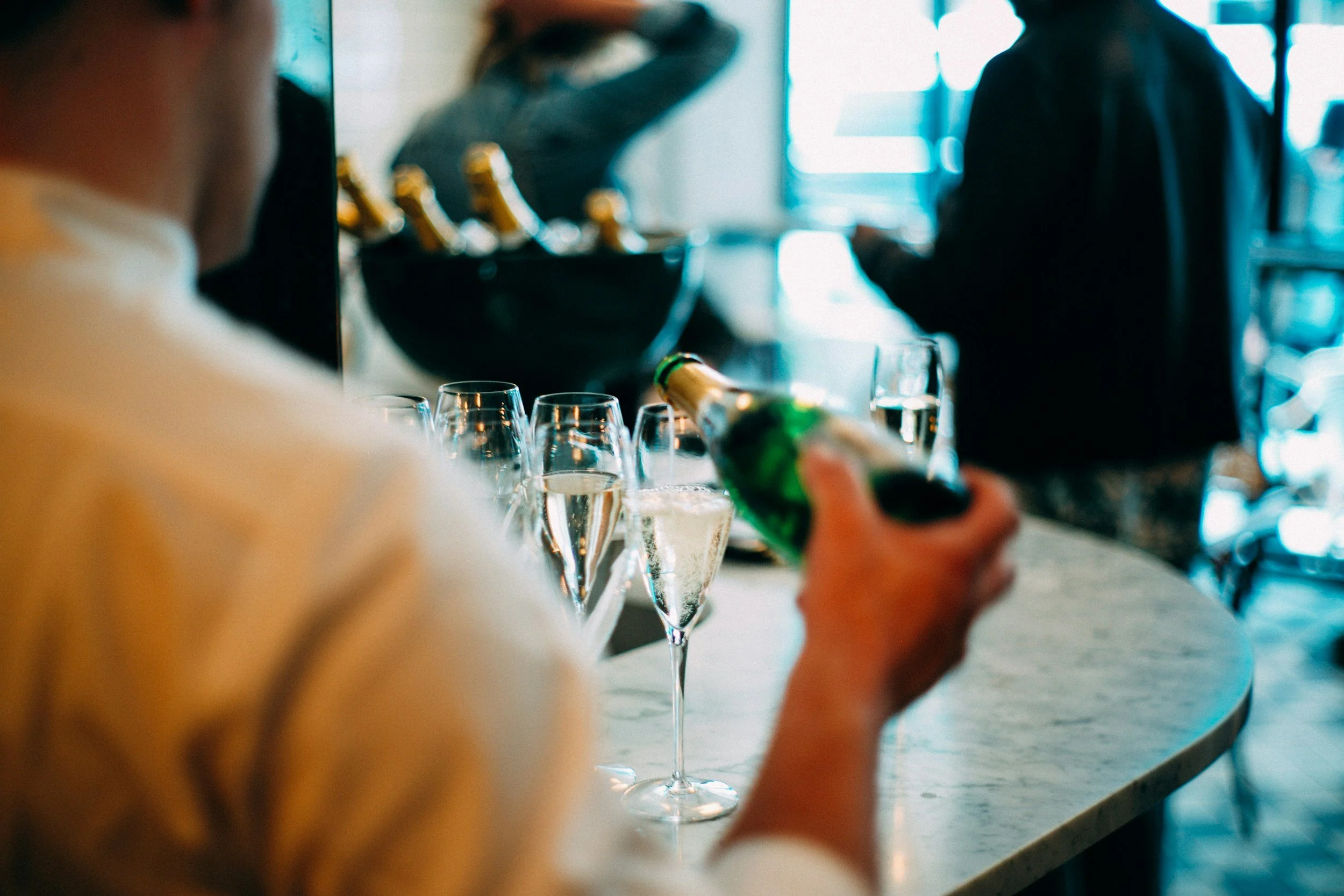 Person pouring champagne into glasses at a bar or party, with others in the background.