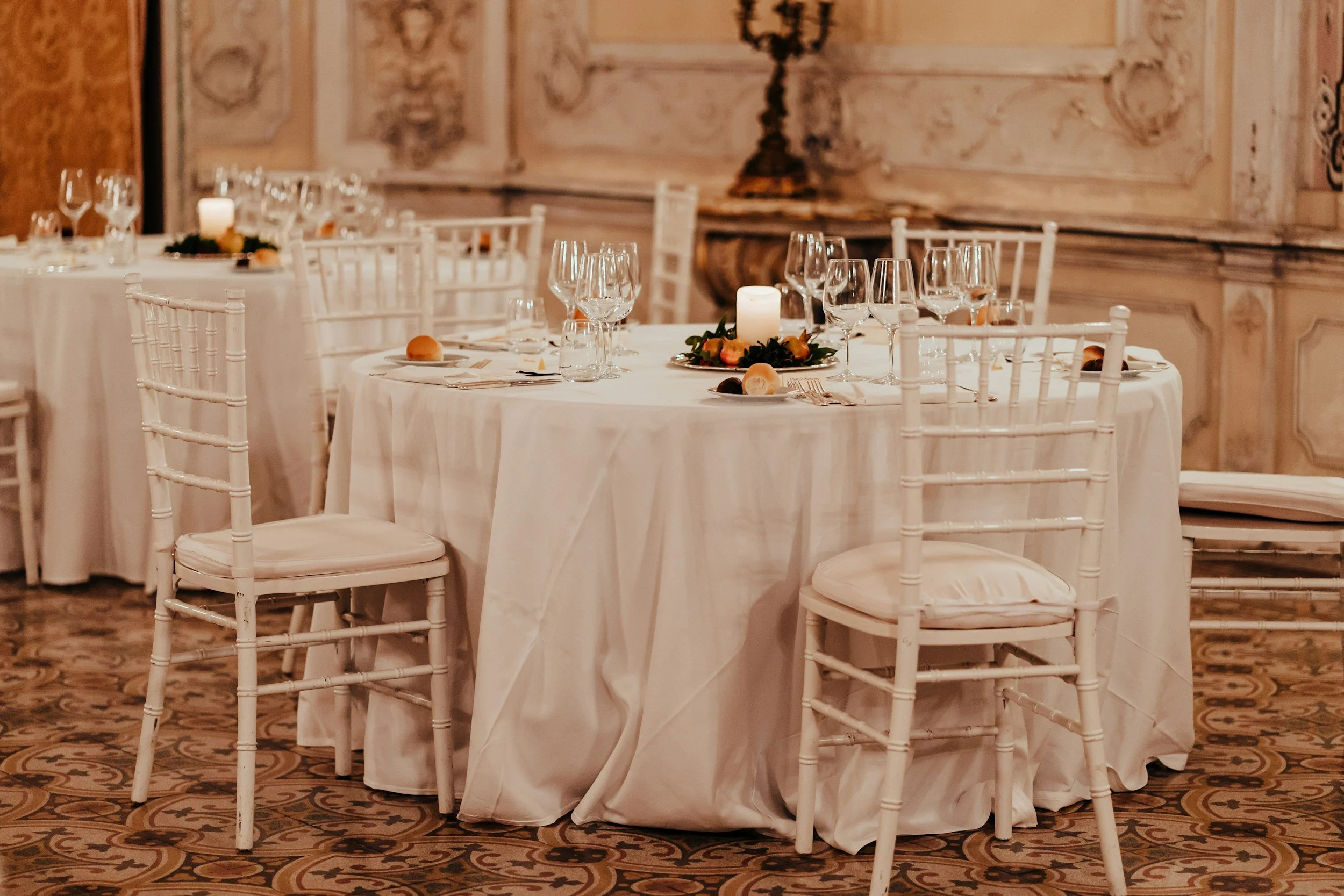 Elegant banquet table set with white tablecloths, multiple wine glasses, glasses of water, silverware, bread rolls, candles, and floral centerpieces, in a decorated room with ornate wall panels and a patterned carpet.