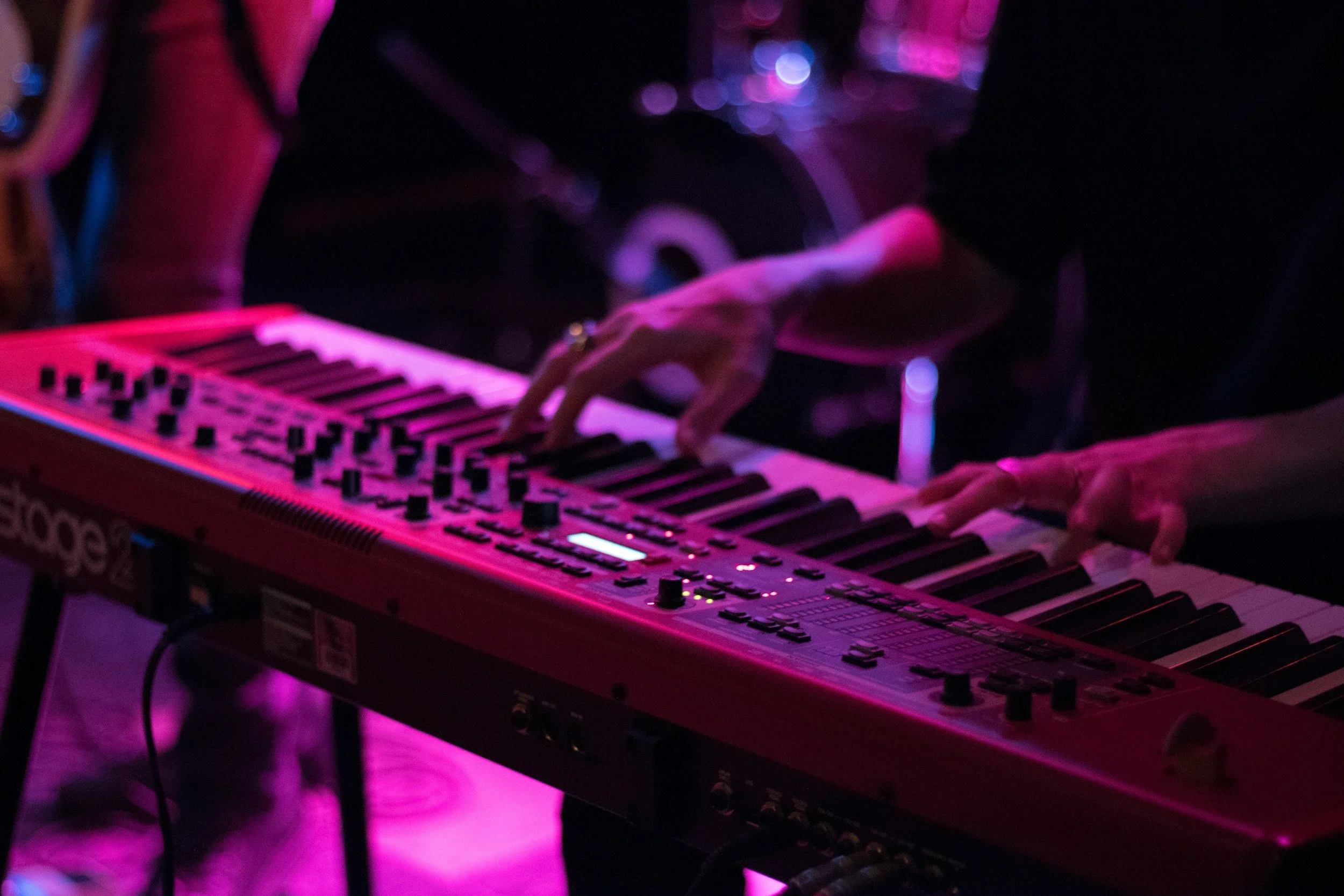A person playing a red keyboard synthesizer in a dimly lit setting with purple and pink lighting.