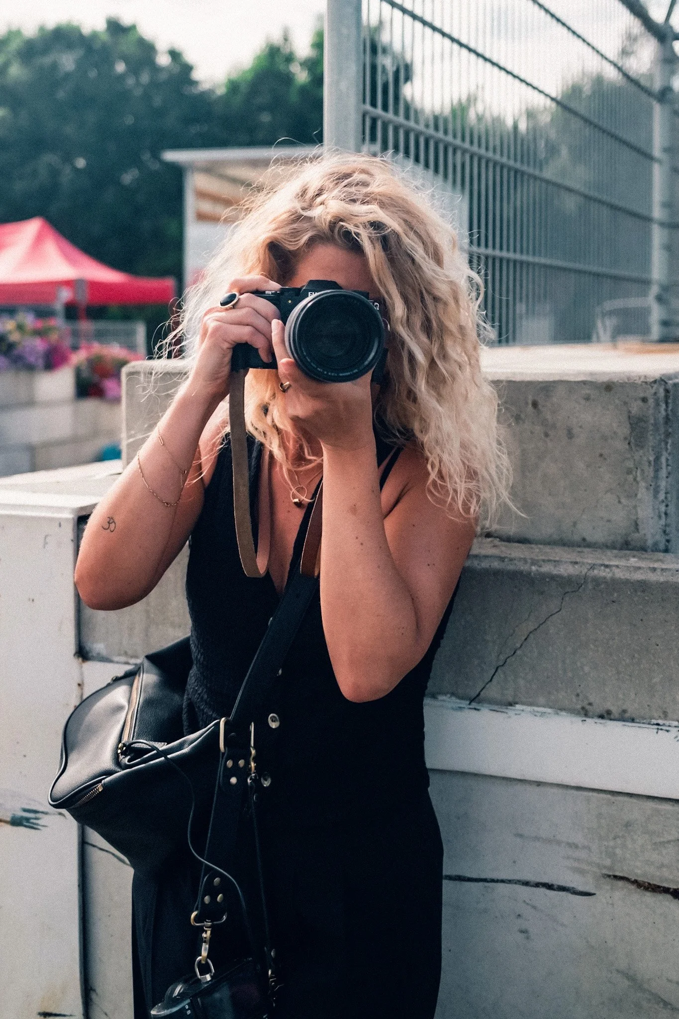 A woman with curly blonde hair taking a photo with a camera outdoors, leaning against a concrete structure near a metal fence, with tents and trees in the background.