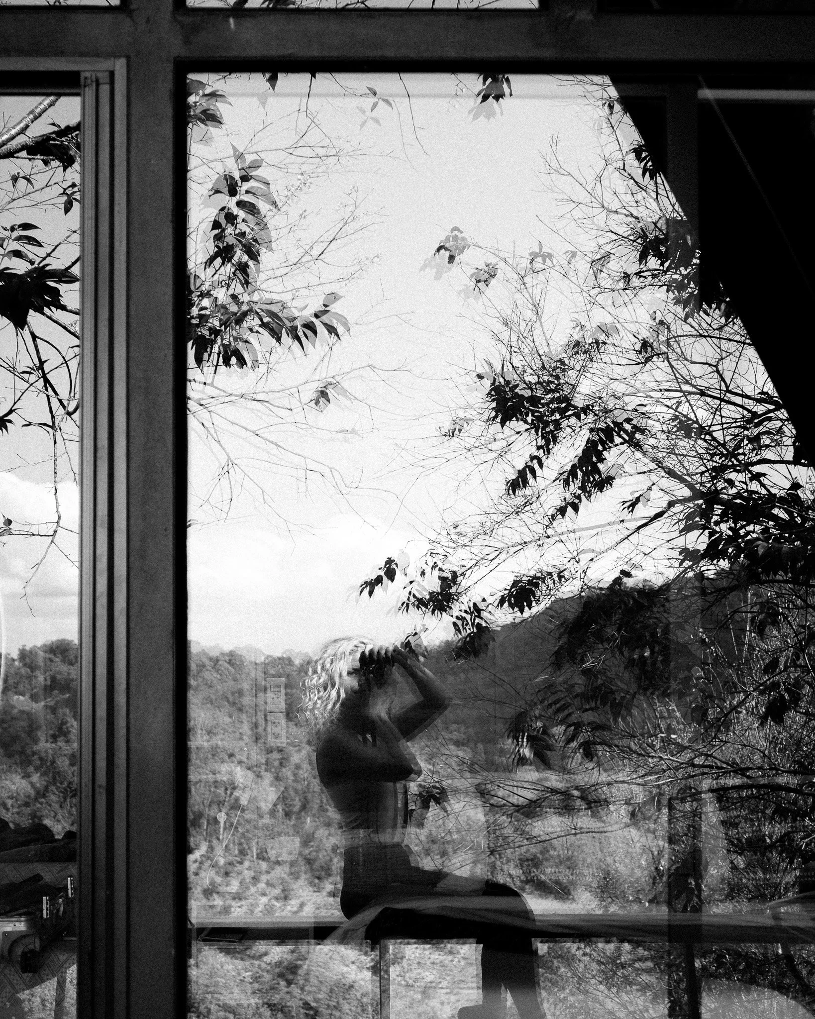 Reflection of a woman with curly hair taking a photo in a window, with trees and a cloudy sky visible outside.