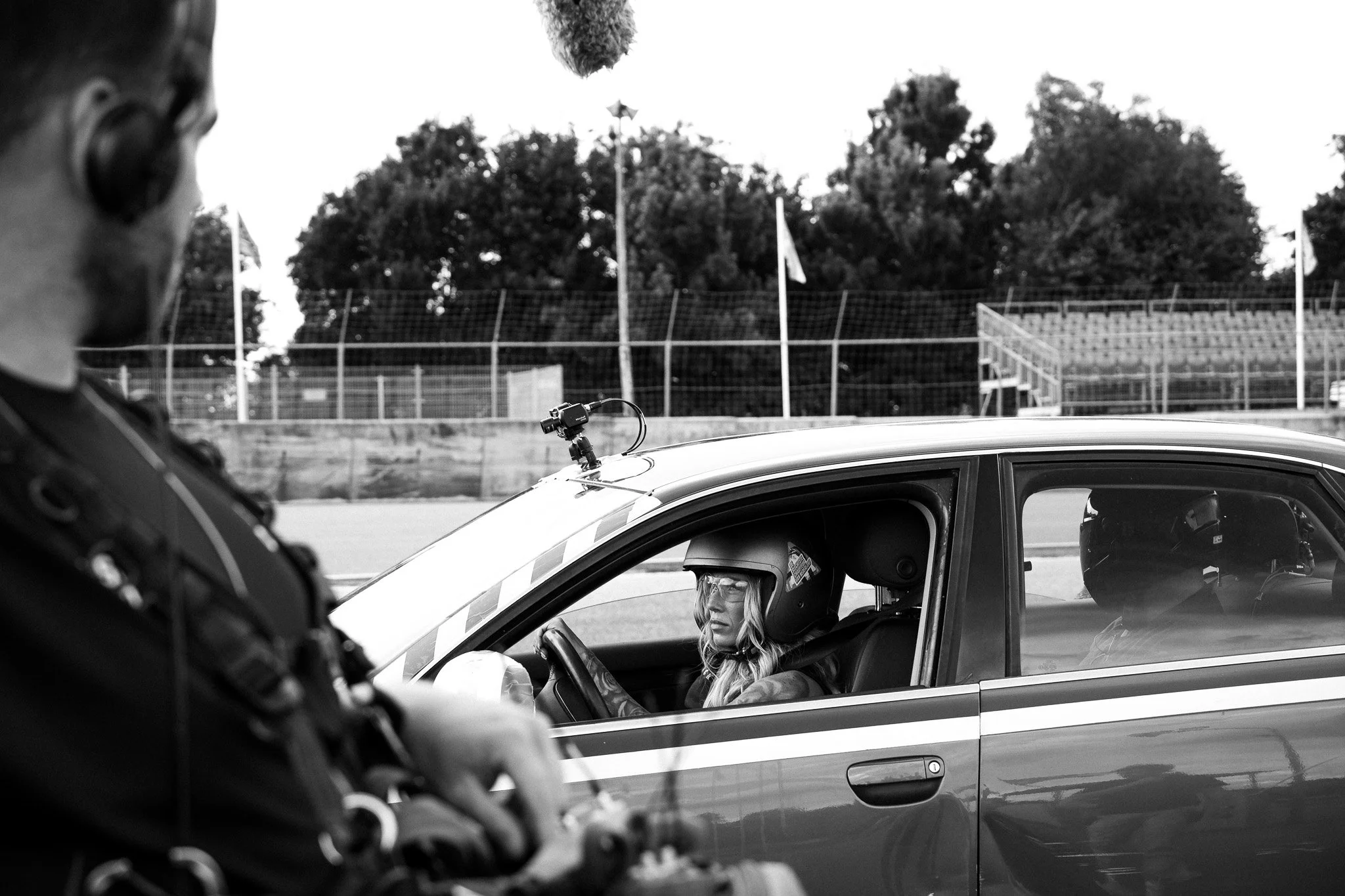 Politieagent in uniform zit in een politiewagen met een vrouwelijke bestuurder op een parkeerplaats, met bomen en een hek op de achtergrond.