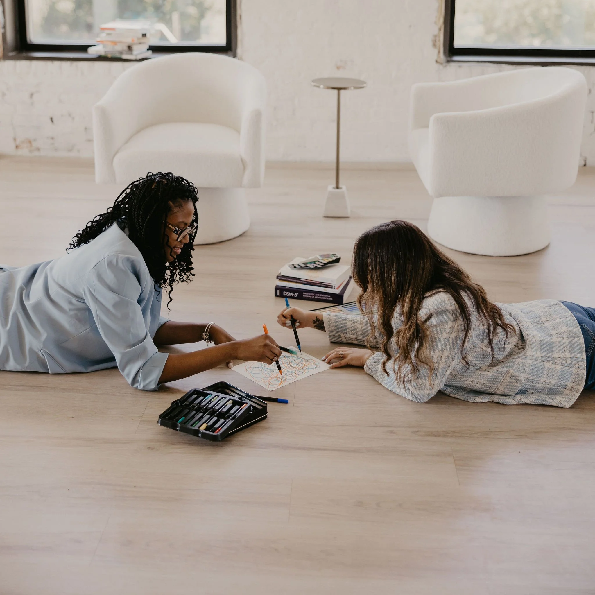 Two women lying on the wooden floor of a modern, minimalist room, drawing with colored markers on paper. The room has two white armchairs, a small table, and books on the floor; large windows with a view of trees.