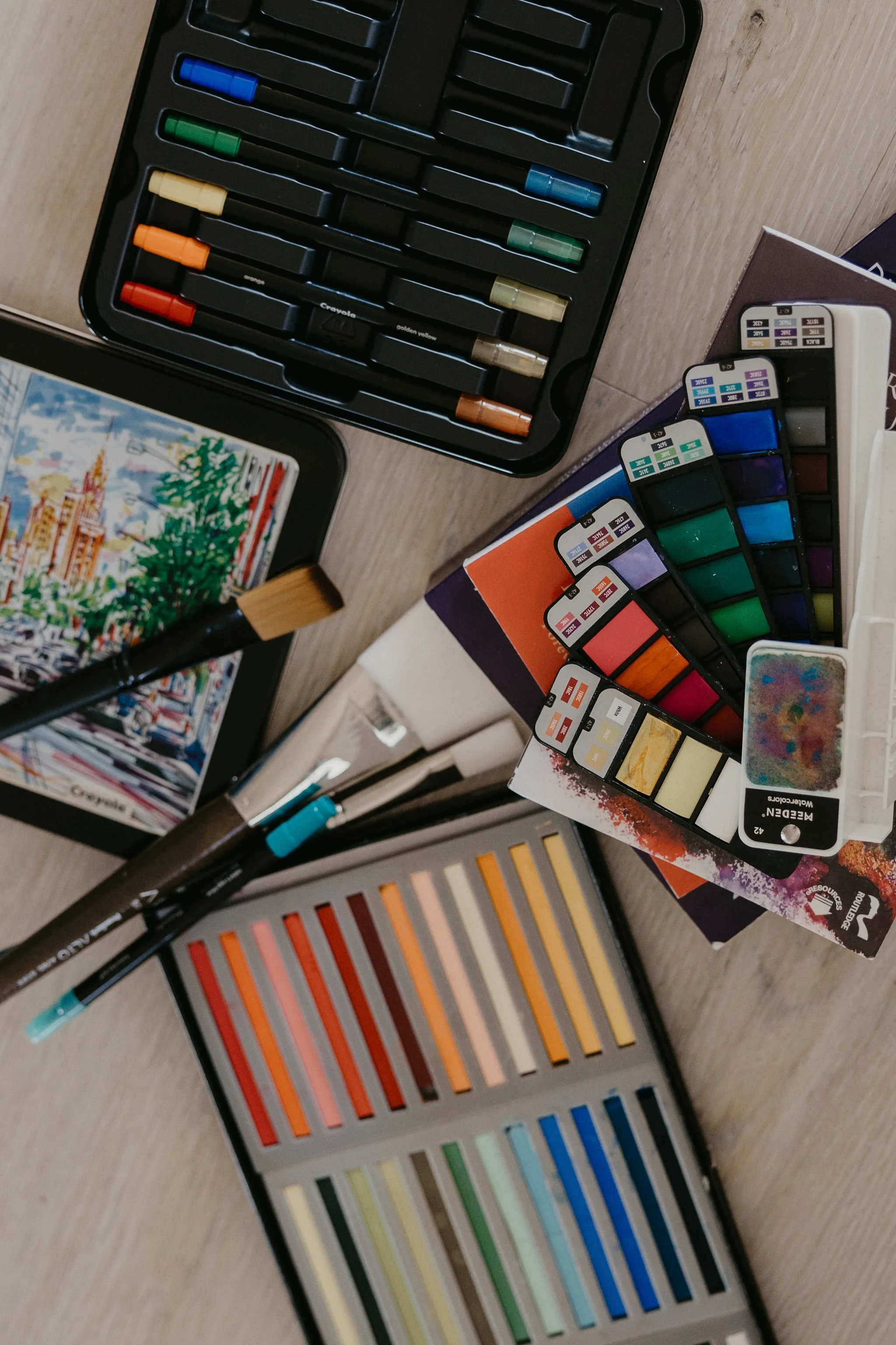 Open watercolor paint set with various colors arranged in rows, paintbrushes around, and color swatches on packaging.