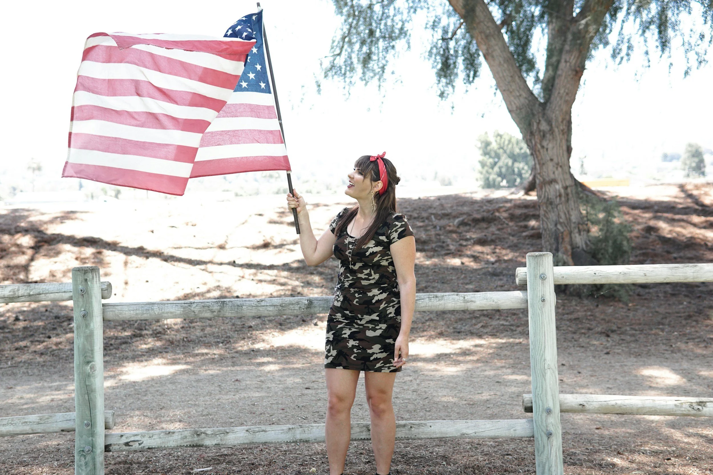 A young woman in a camouflage dress holding an American flag outdoors near a wooden fence, with trees and sky in the background.