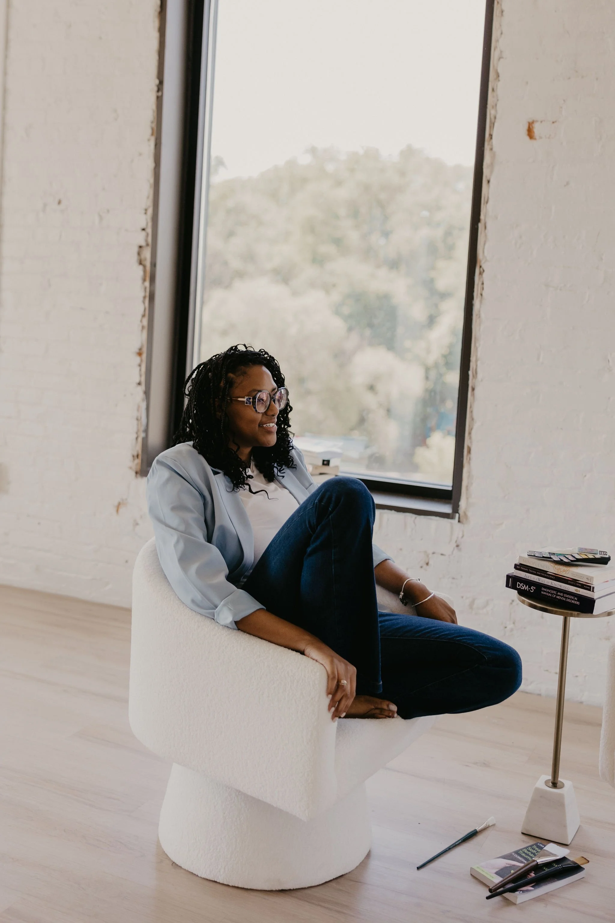 A woman with glasses and curly hair sitting on a white modern chair by a large window in a minimalist room, reading a book.