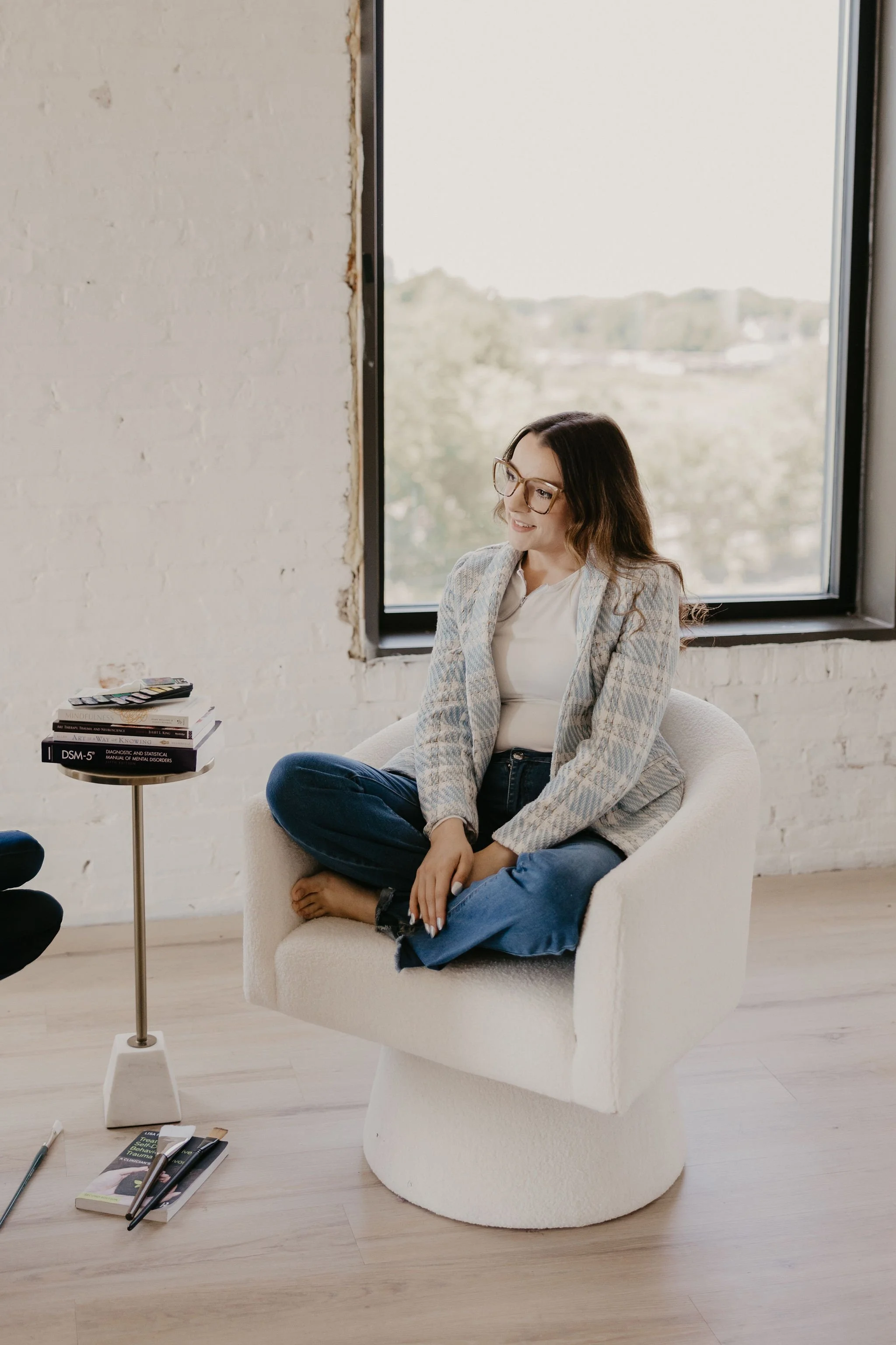 A woman with glasses and long hair, sitting cross-legged on a white modern chair, smiling, in a room with a large window, a small table with books, and a white brick wall.