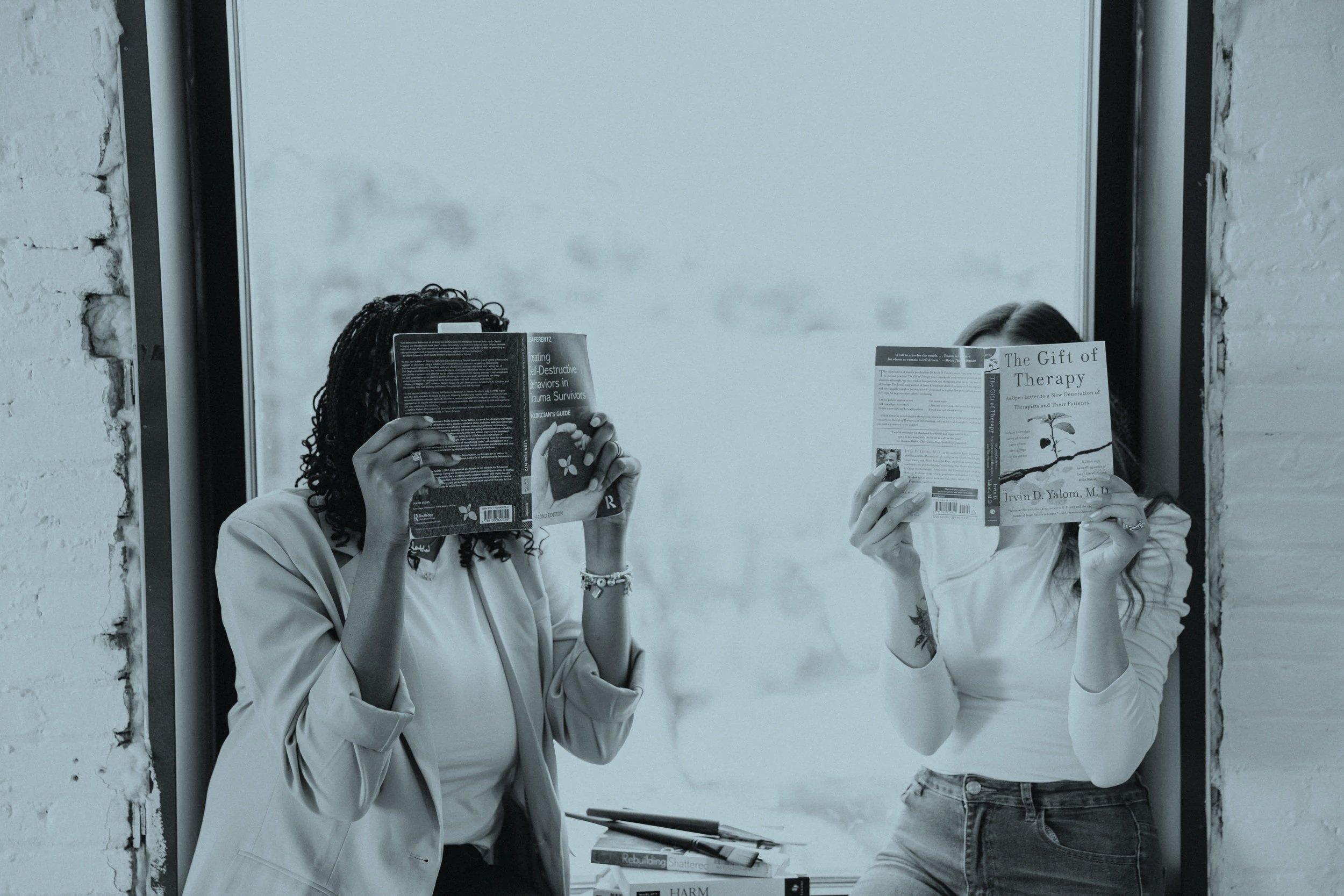 Two women standing by a window, reading books, with a stack of books on a table between them.