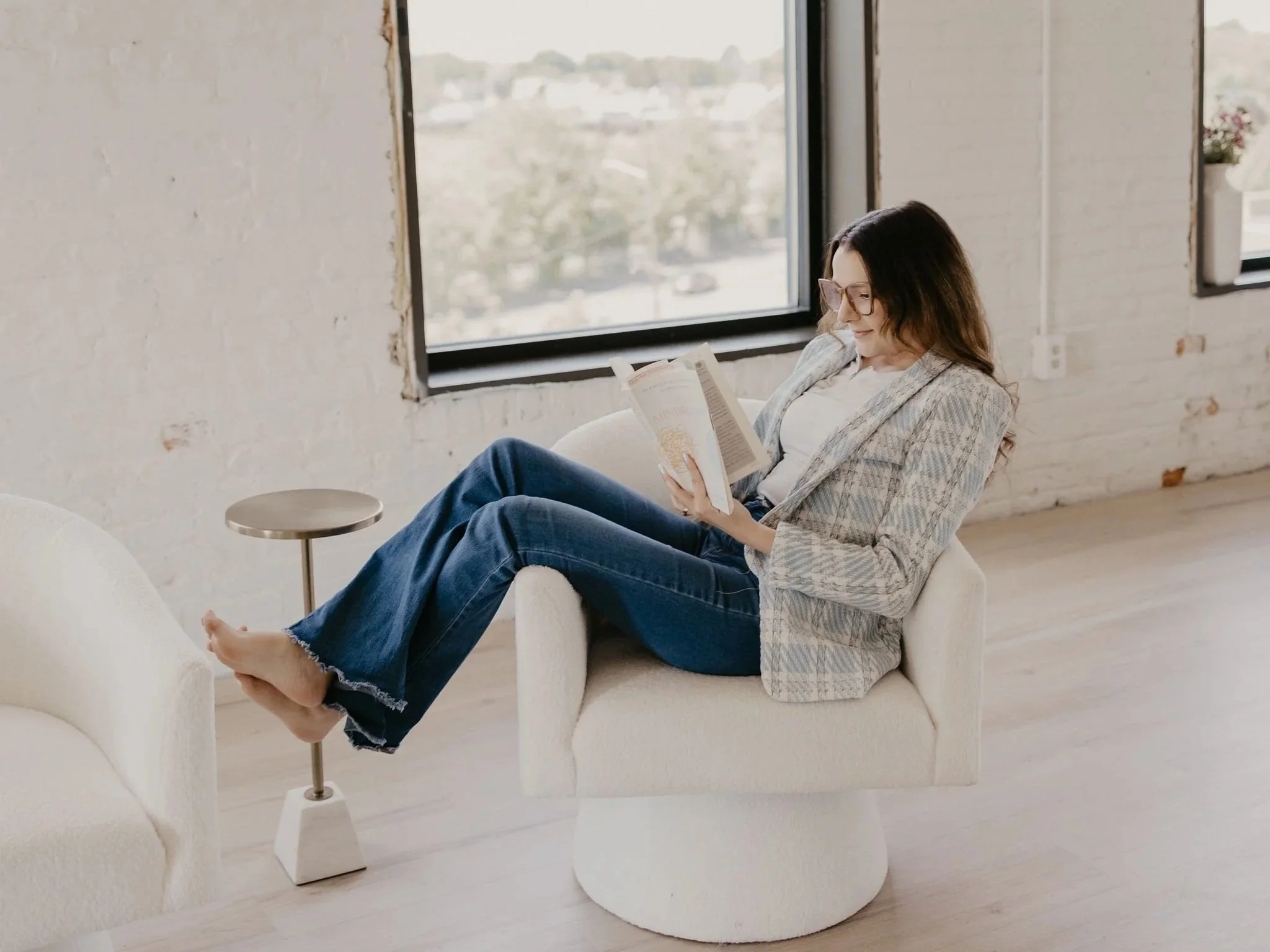 A woman with glasses, wearing a plaid blazer and jeans, sitting barefoot in an off-white armchair, reading a book with her legs extended across a small side table, in a bright room with large windows and a white brick wall.