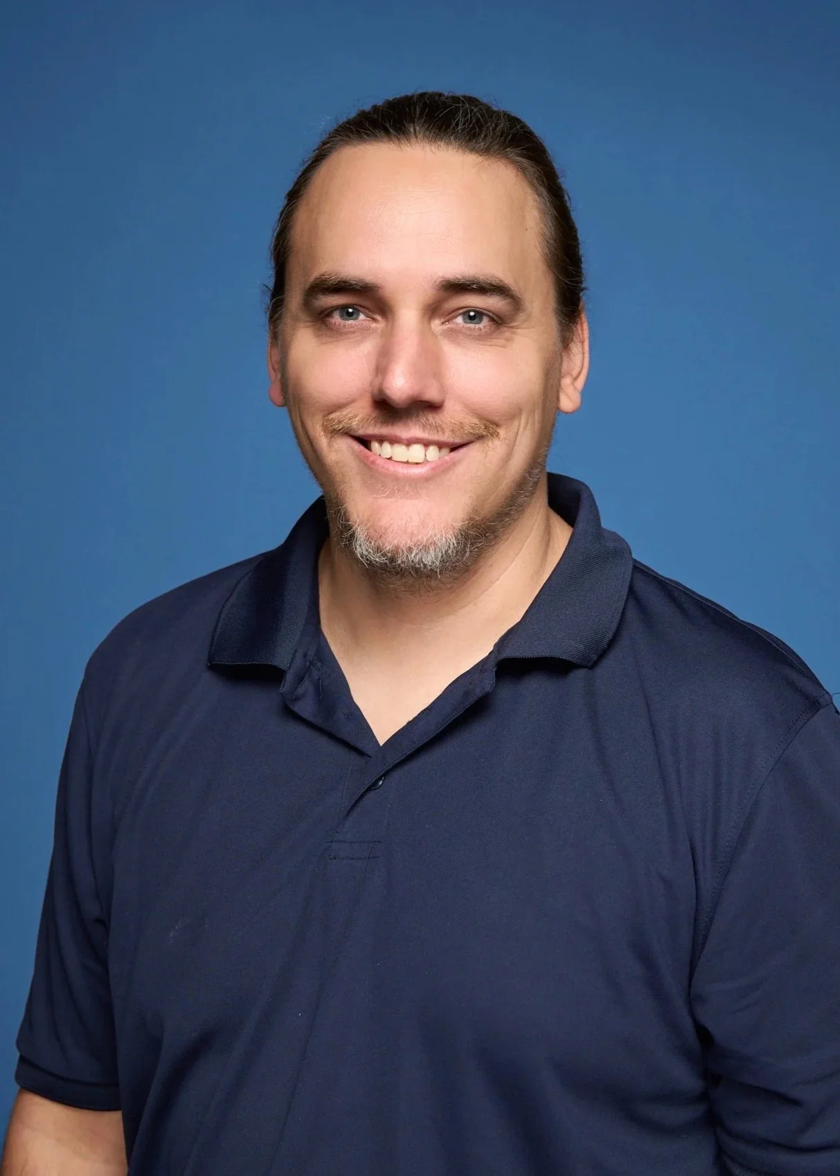 Headshot of a smiling man with light skin, long brown hair tied back, and a goatee, wearing a navy blue polo shirt against a solid blue background.