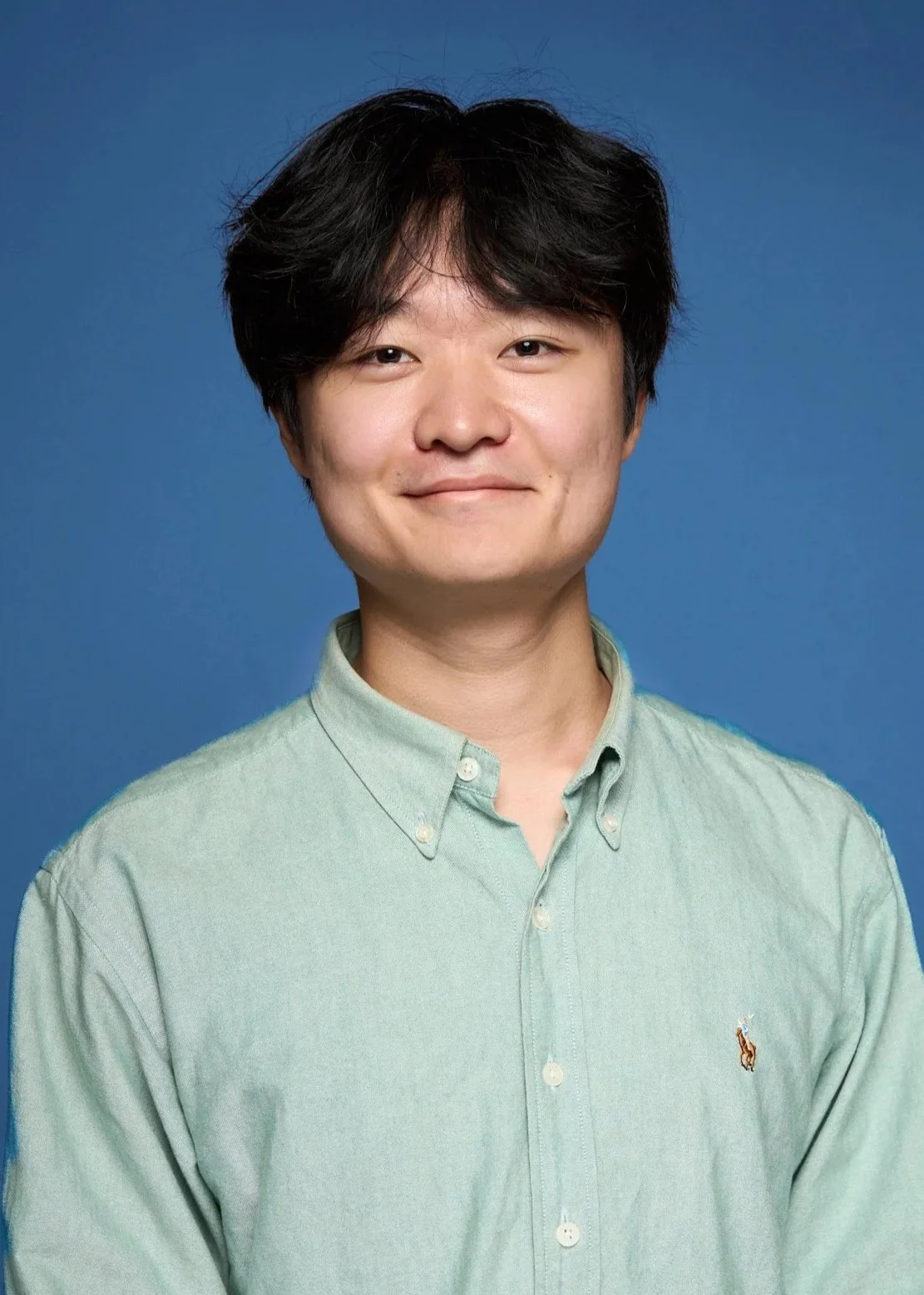 A young man with dark hair smiling, wearing a light green button-up shirt with a small embroidered logo, against a blue background.