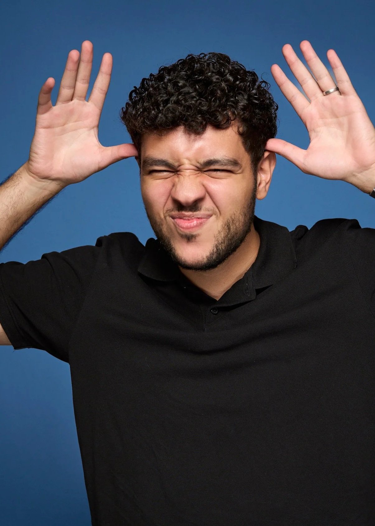 A man with curly dark hair and a beard making a face with eyes closed, squinting, and mouth slightly scrunched, wearing a black polo shirt and holding his hands near his temples with fingers spread out against a blue background.