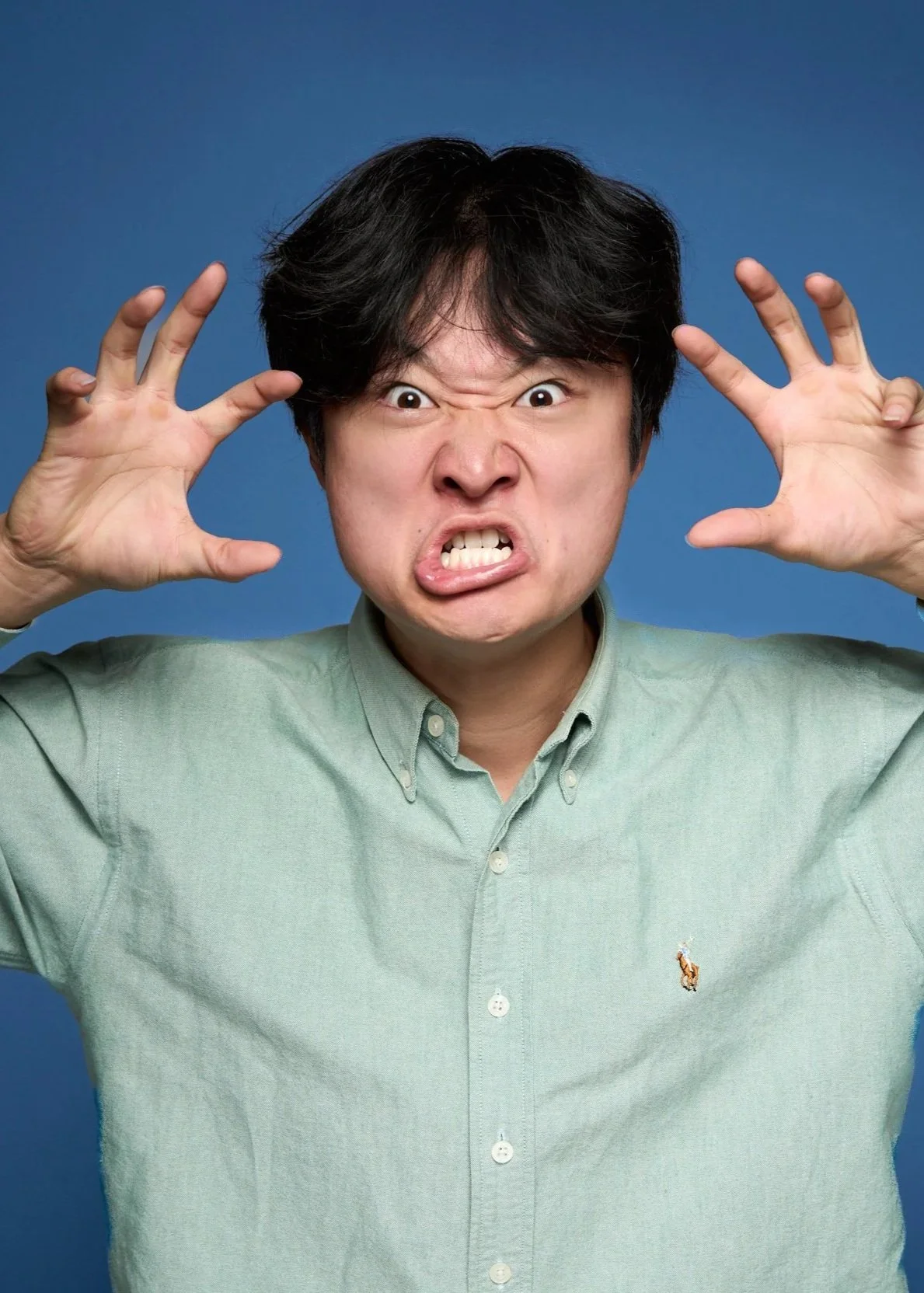 Man with black hair making an angry face, hands near his head, against a blue background.