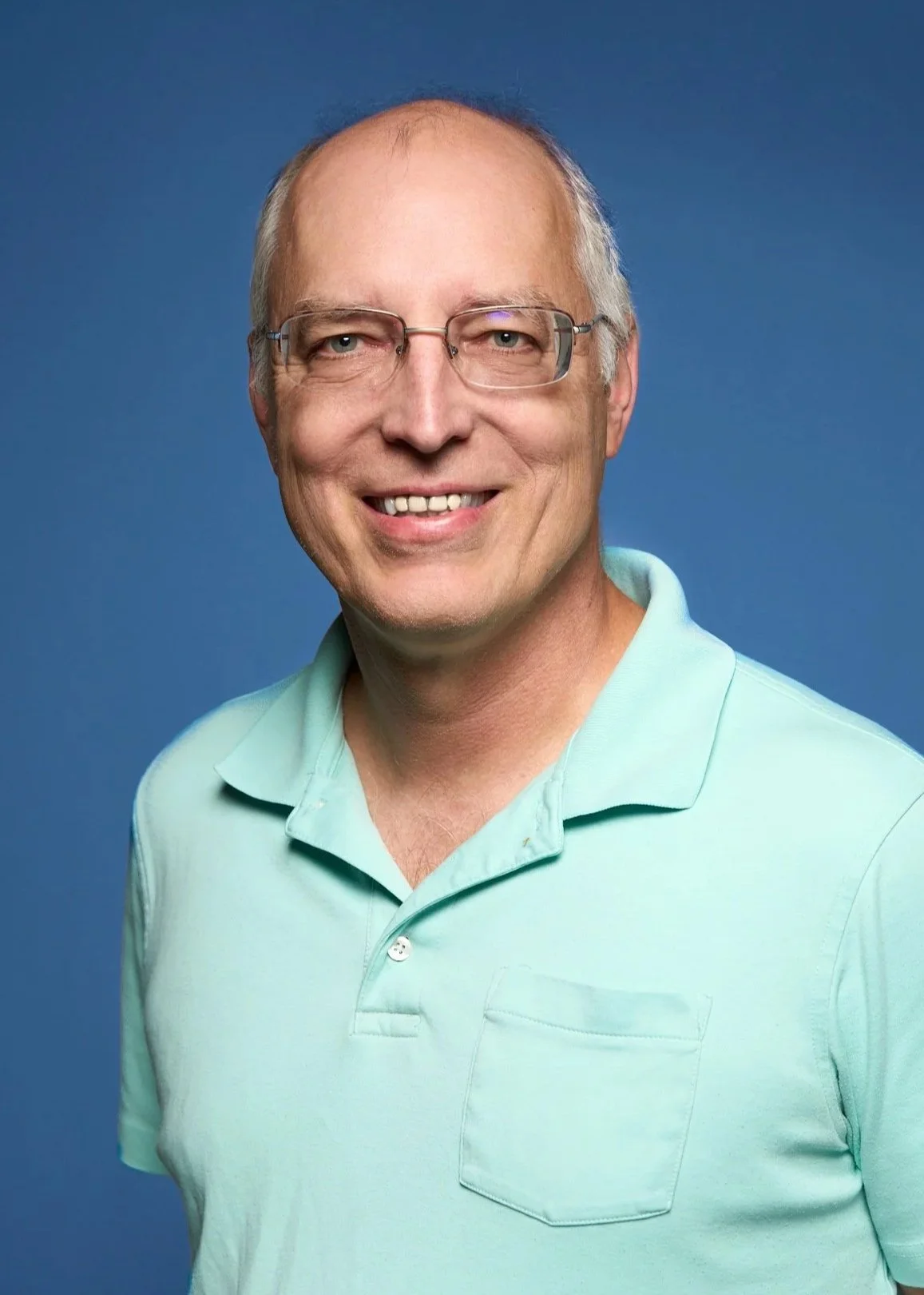 Portrait of a middle-aged man with glasses, smiling, wearing a light green polo shirt, against a blue background.