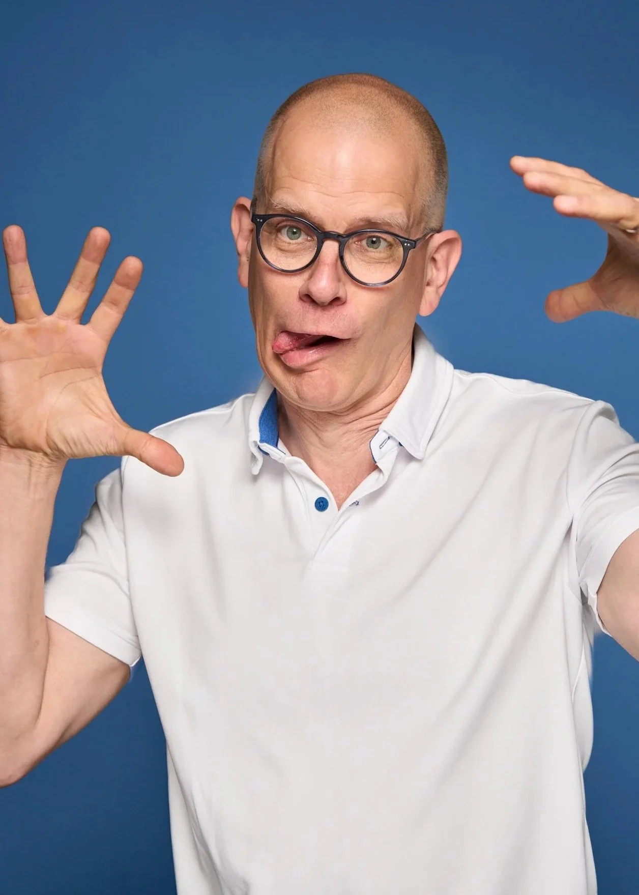 Man with glasses making silly face, tongue out, hands raised, against a blue background.
