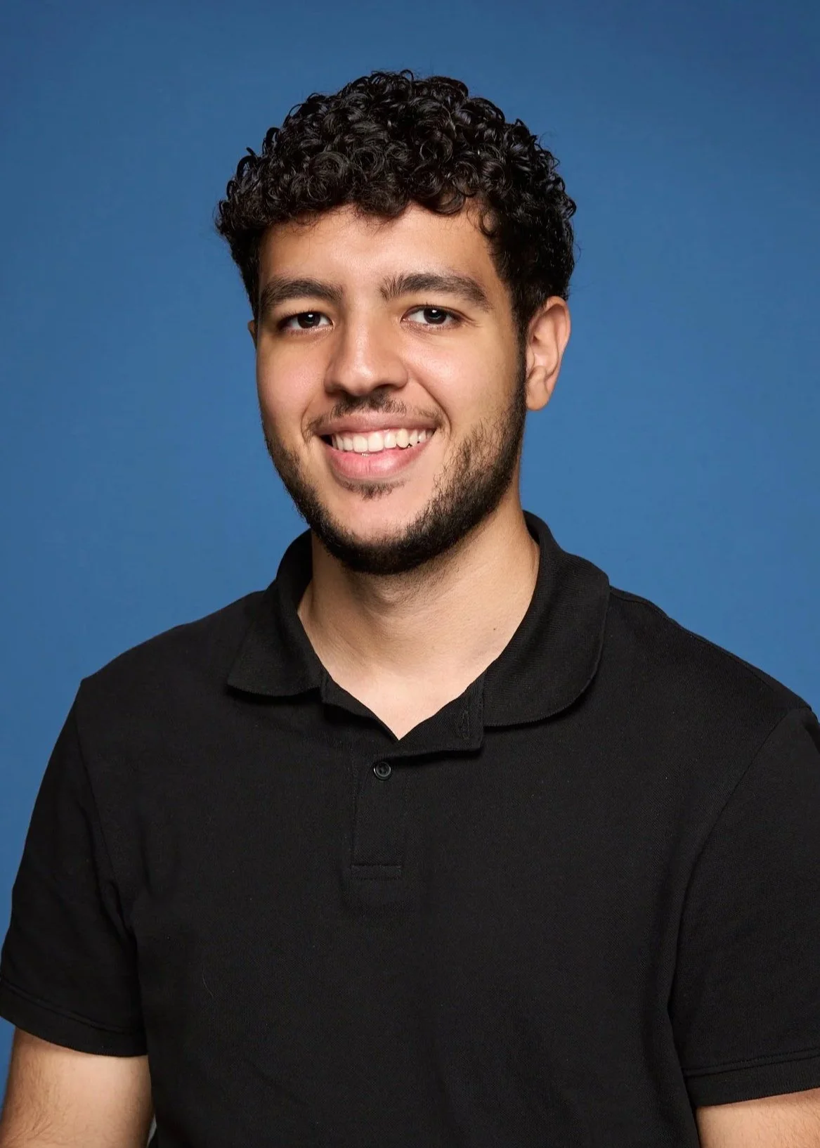 A young man with curly dark hair, a beard, and a mustache, smiling, wearing a black collared shirt, against a solid blue background.