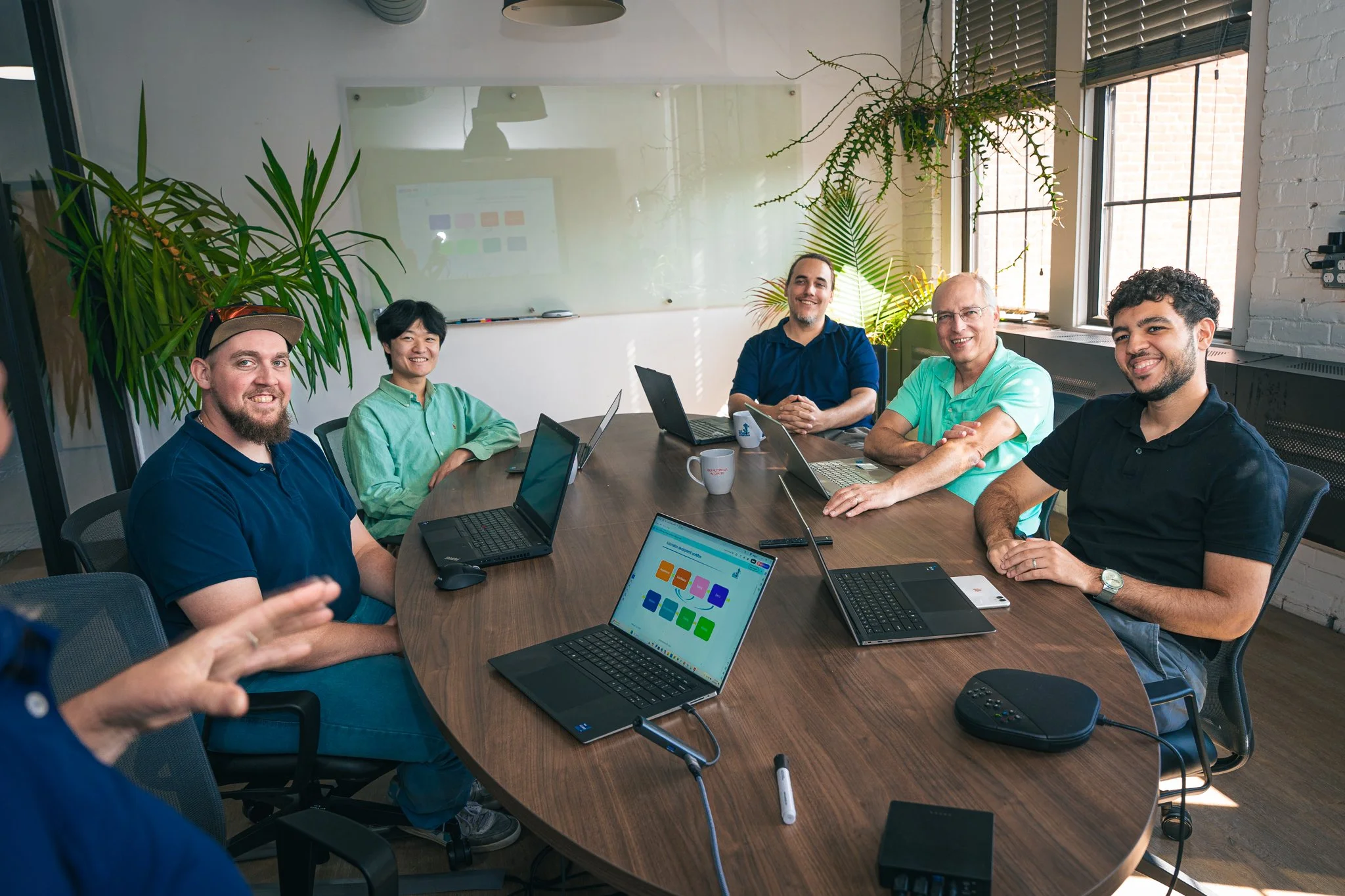 Group of six people in a meeting room, sitting around a large oval table with laptops, mugs, and conference call equipment, smiling and engaging with each other.